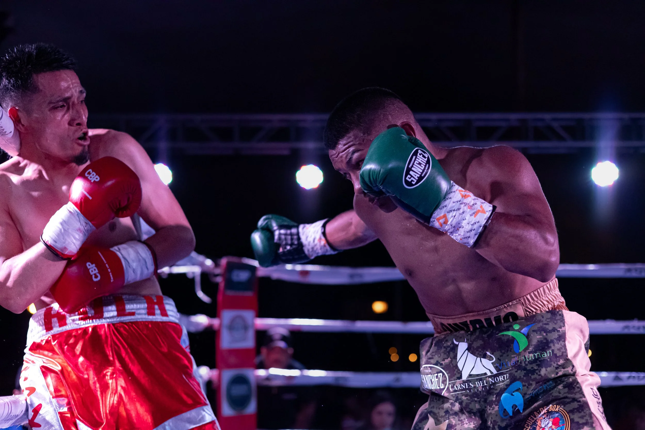 Two shirtless male boxers fighting in a boxing ring at night, one wearing red gloves and shorts, the other wearing green gloves and camouflage shorts, with bright lights overhead.