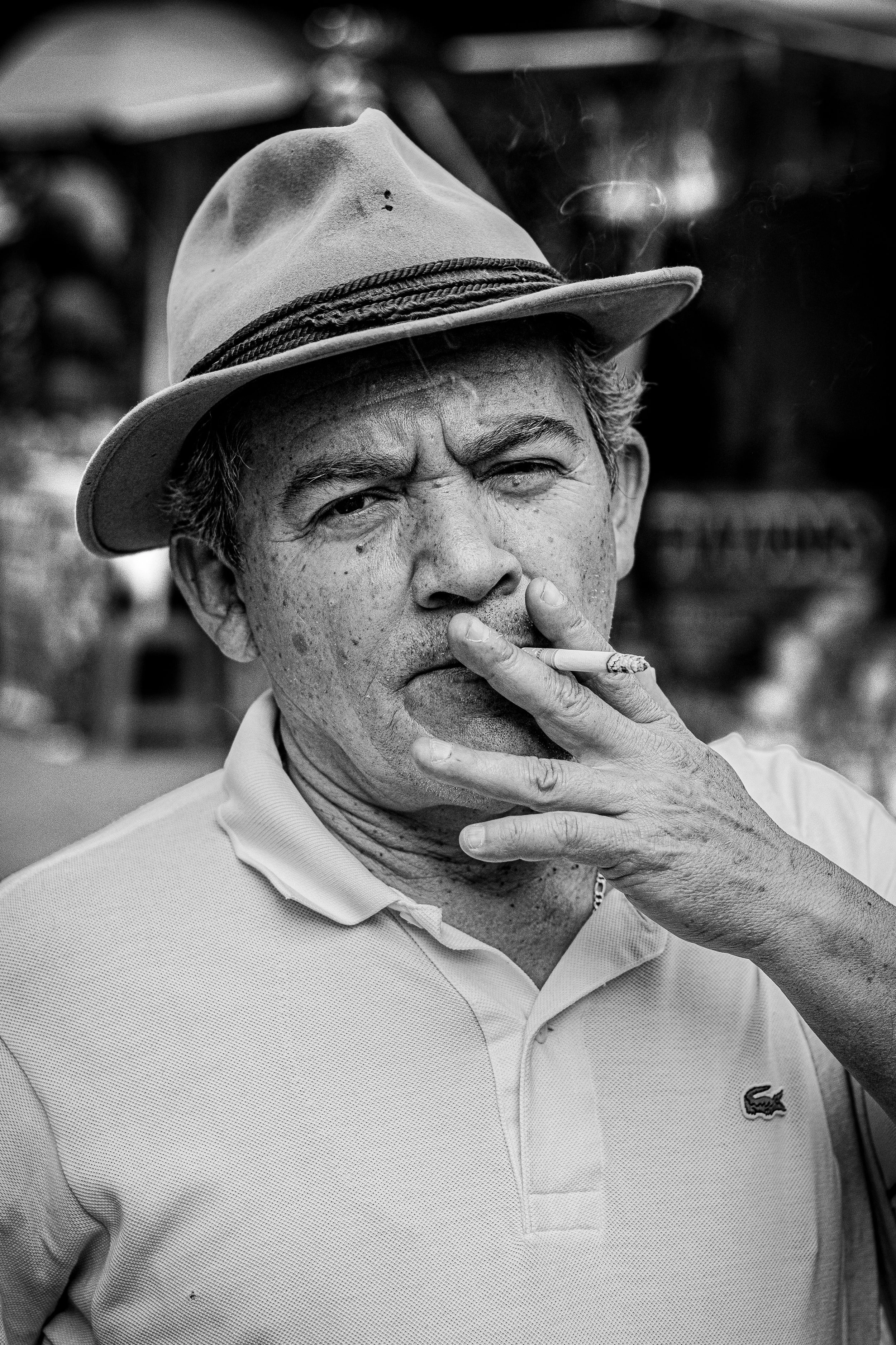 Black and white photo of an older man smoking a cigarette, wearing a hat and a polo shirt.