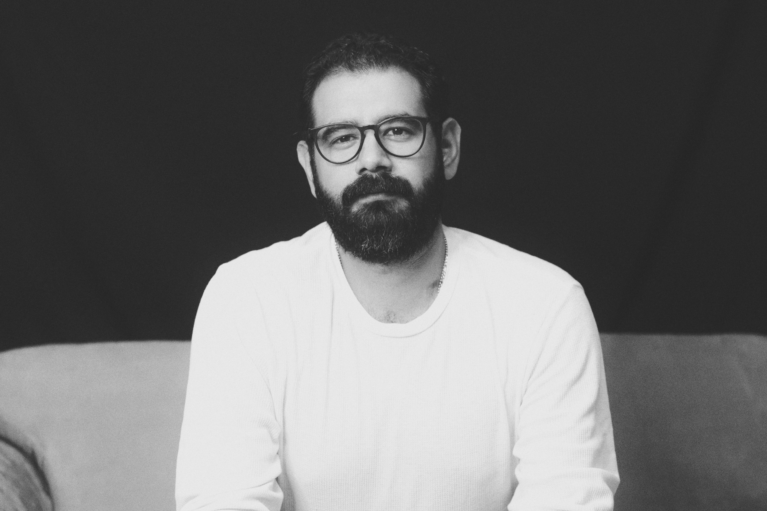 A man with glasses, a full beard, and short hair, wearing a white shirt, sitting on a sofa against a dark background.