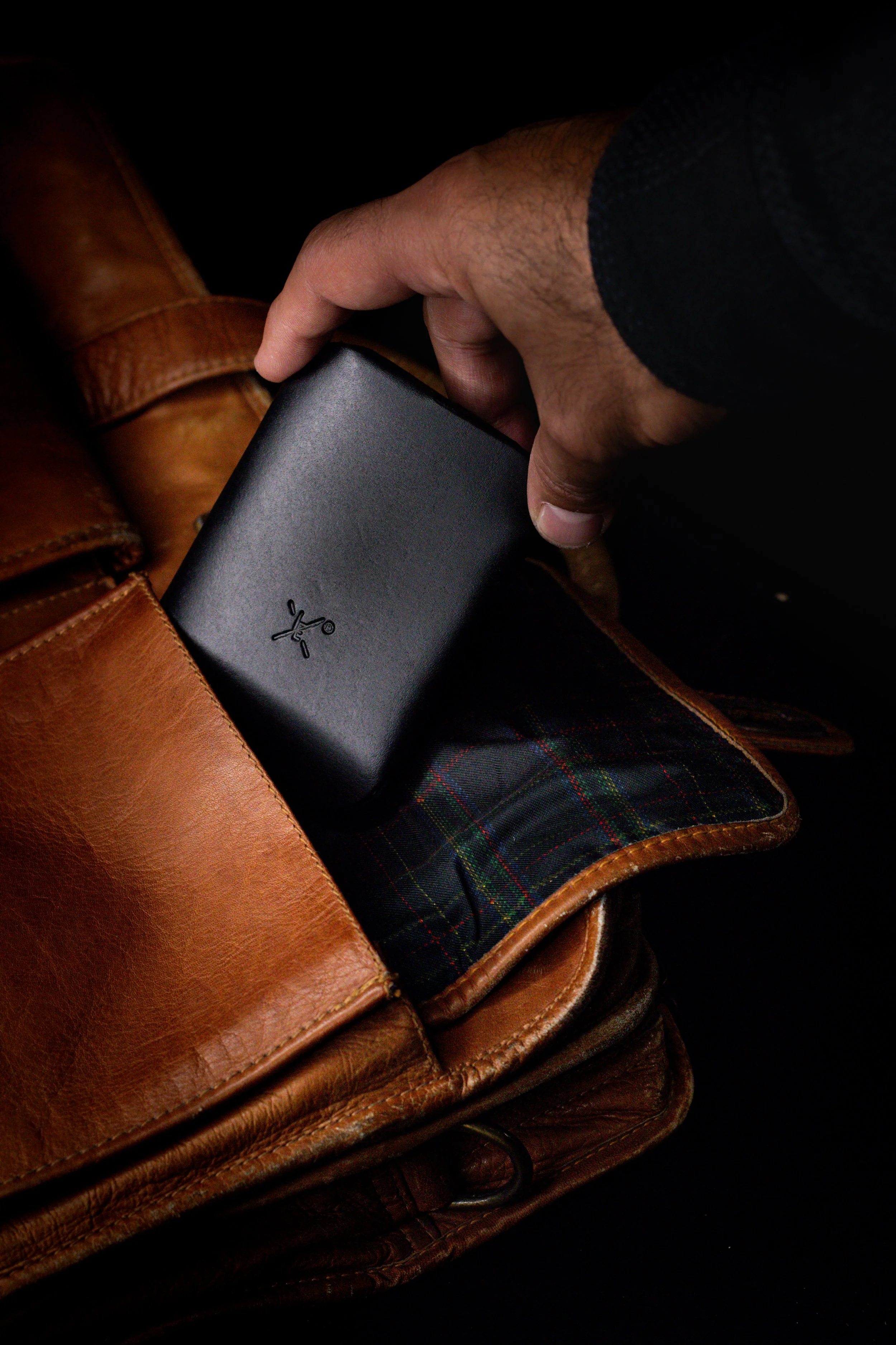 A hand placing a black wireless charger into a leather bag with a tartan-patterned interior.