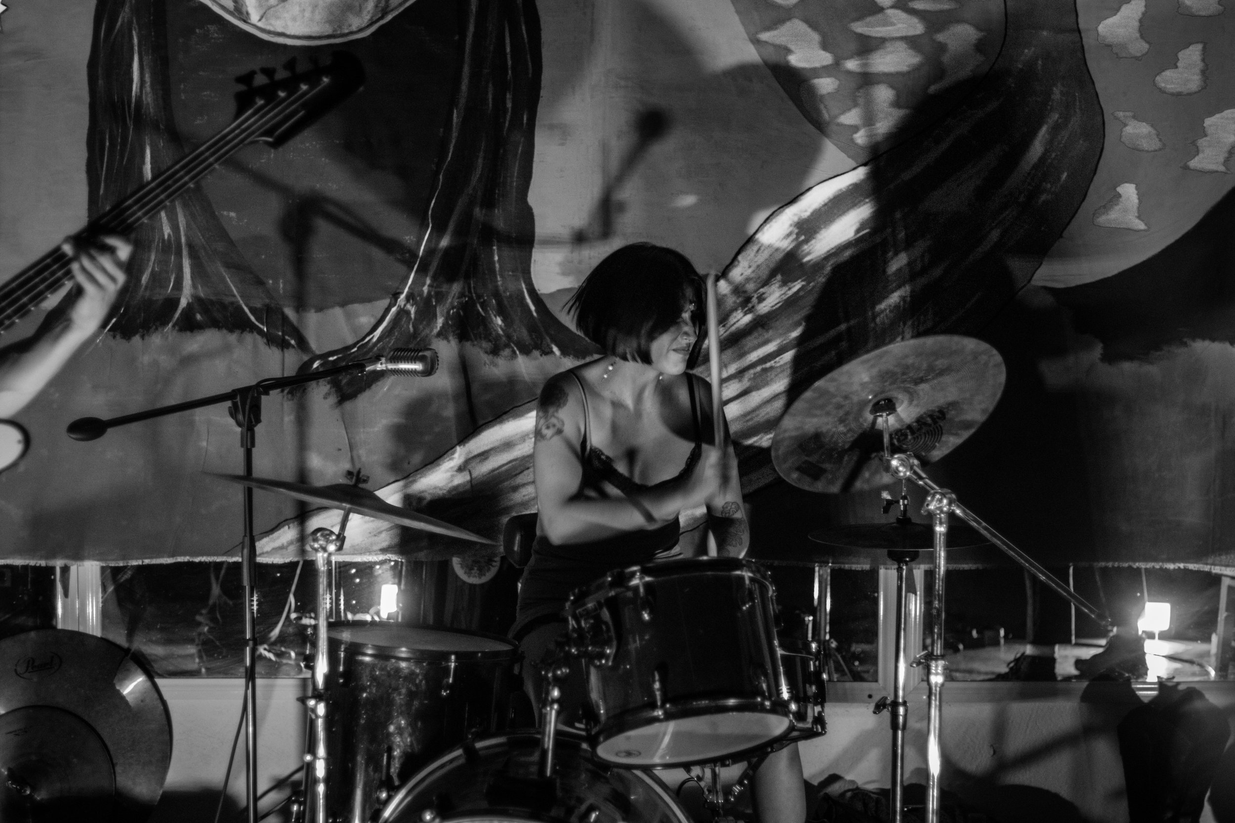 A woman playing drums during a live music performance in a dimly lit venue with a colorful abstract mural in the background.