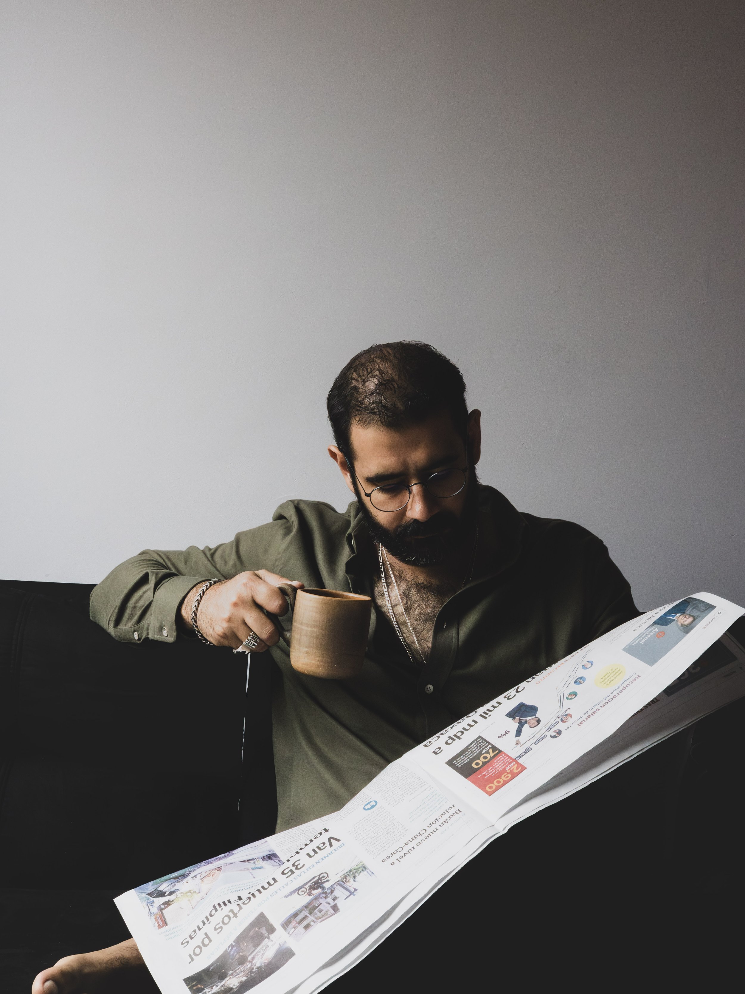 A man with glasses and a beard is sitting at a table, reading a newspaper and holding a ceramic mug.