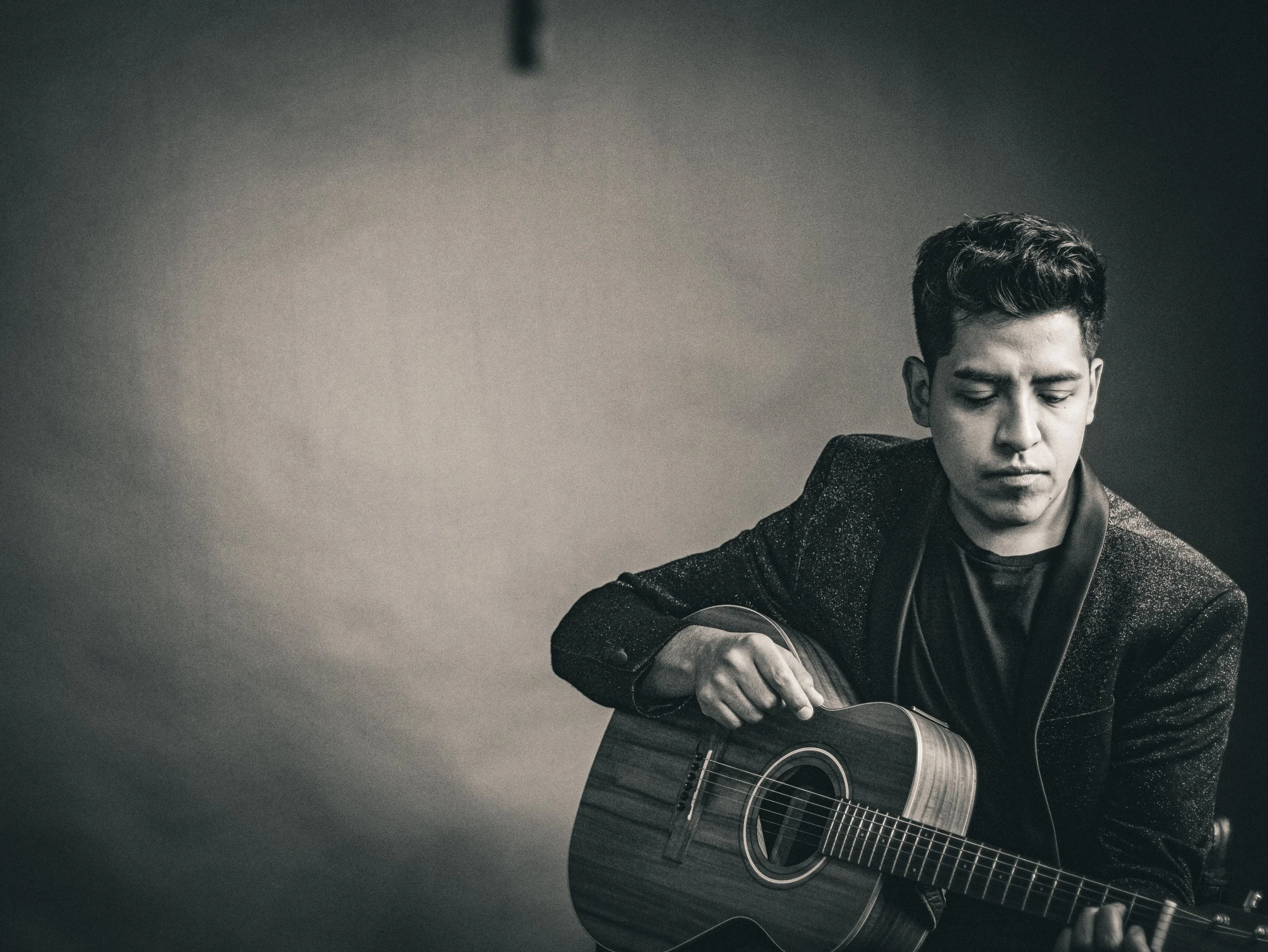 A young man playing an acoustic guitar, looking down in a contemplative mood, black and white image.
