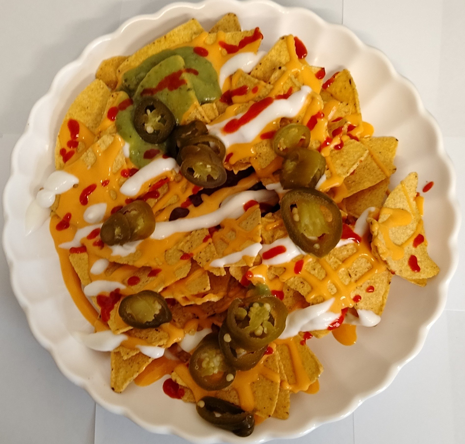 Plate of nachos with tortilla chips, cheese sauce, sour cream, jalapeño slices, red sauce, and shredded cheese.