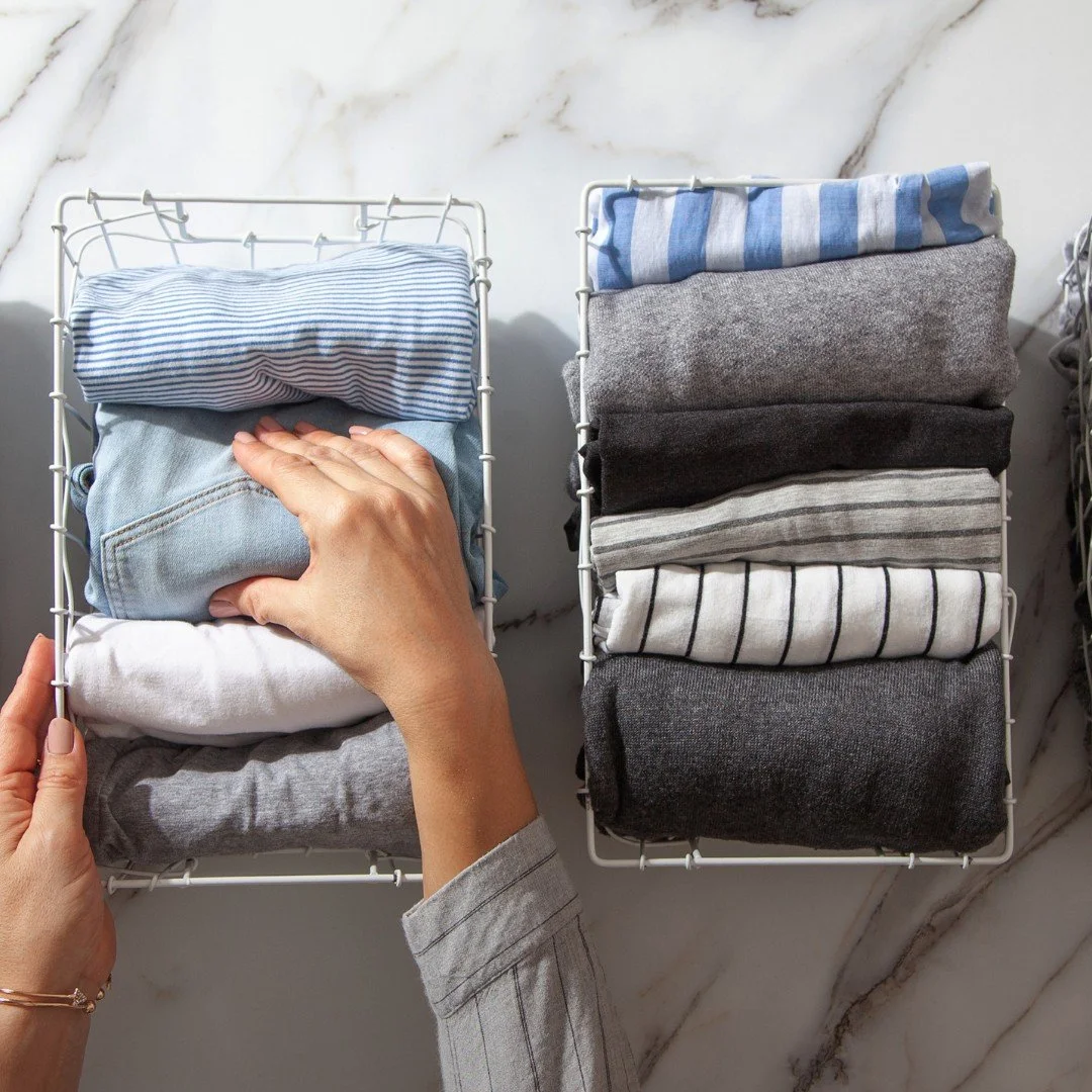 Hands folding T-shirts and sweaters into wire baskets on a marble counter