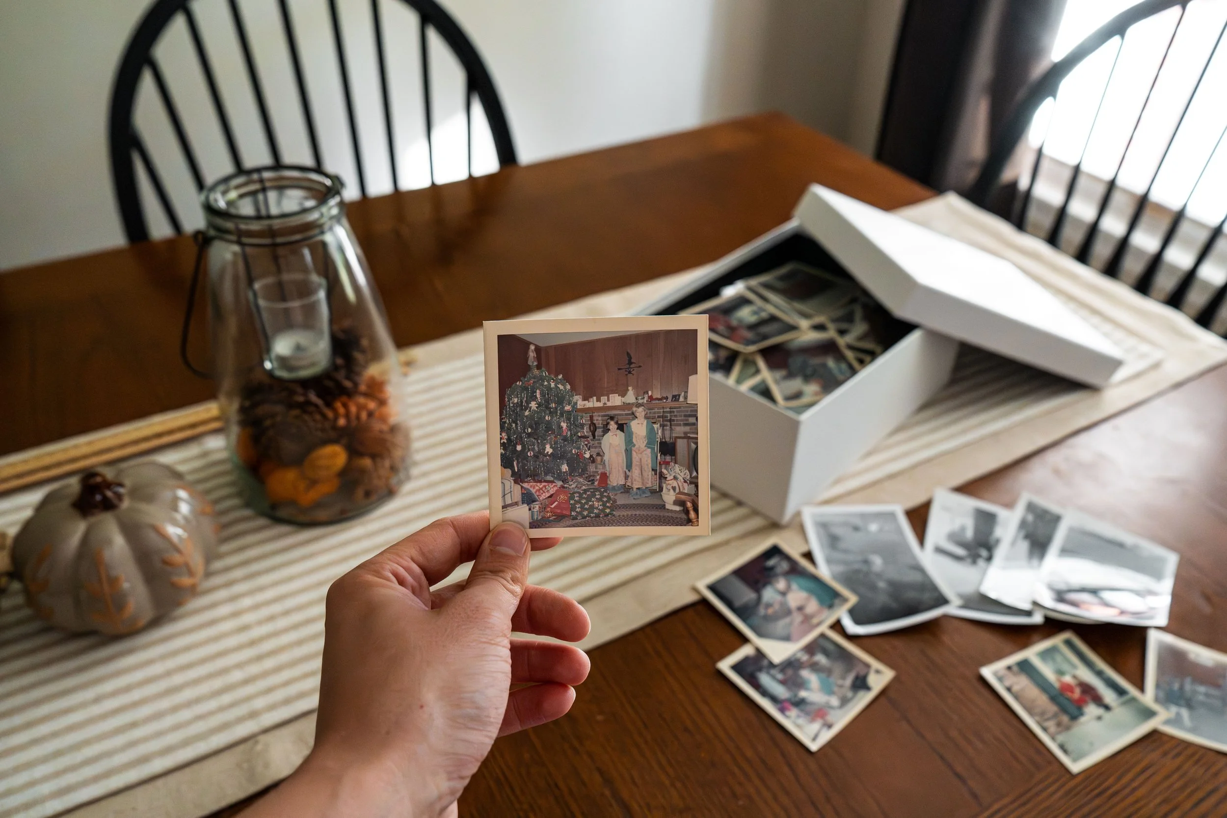 A hand holding a vintage Christmas photo of two children in bathrobes beside a decorated tree, with a box of old family photos on a dining table while preparing a home for a move