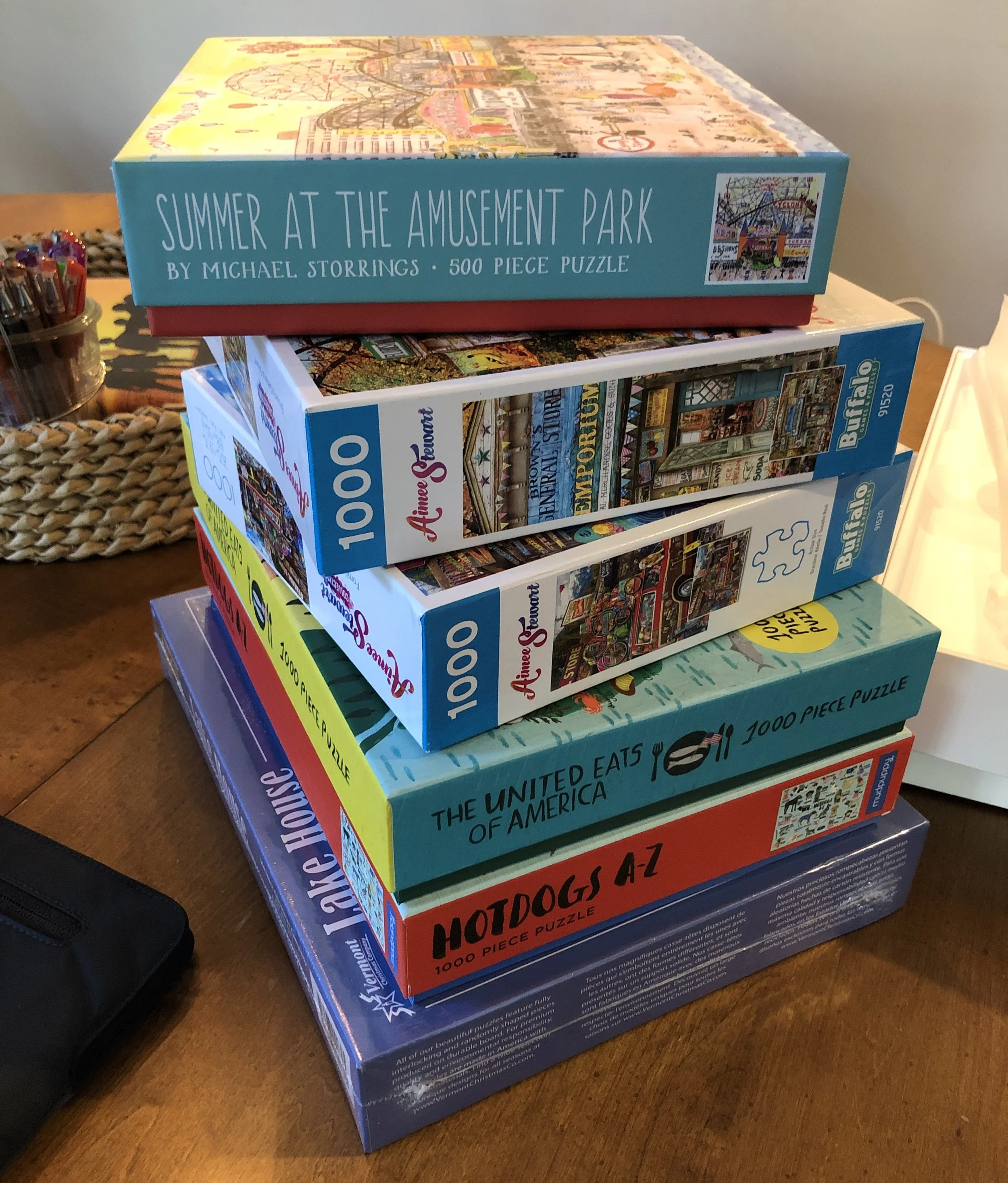 A tall stack of boxed jigsaw puzzles on a wooden table, including Summer at the Amusement Park, The United Eats of America, and Hotdogs A-Z.