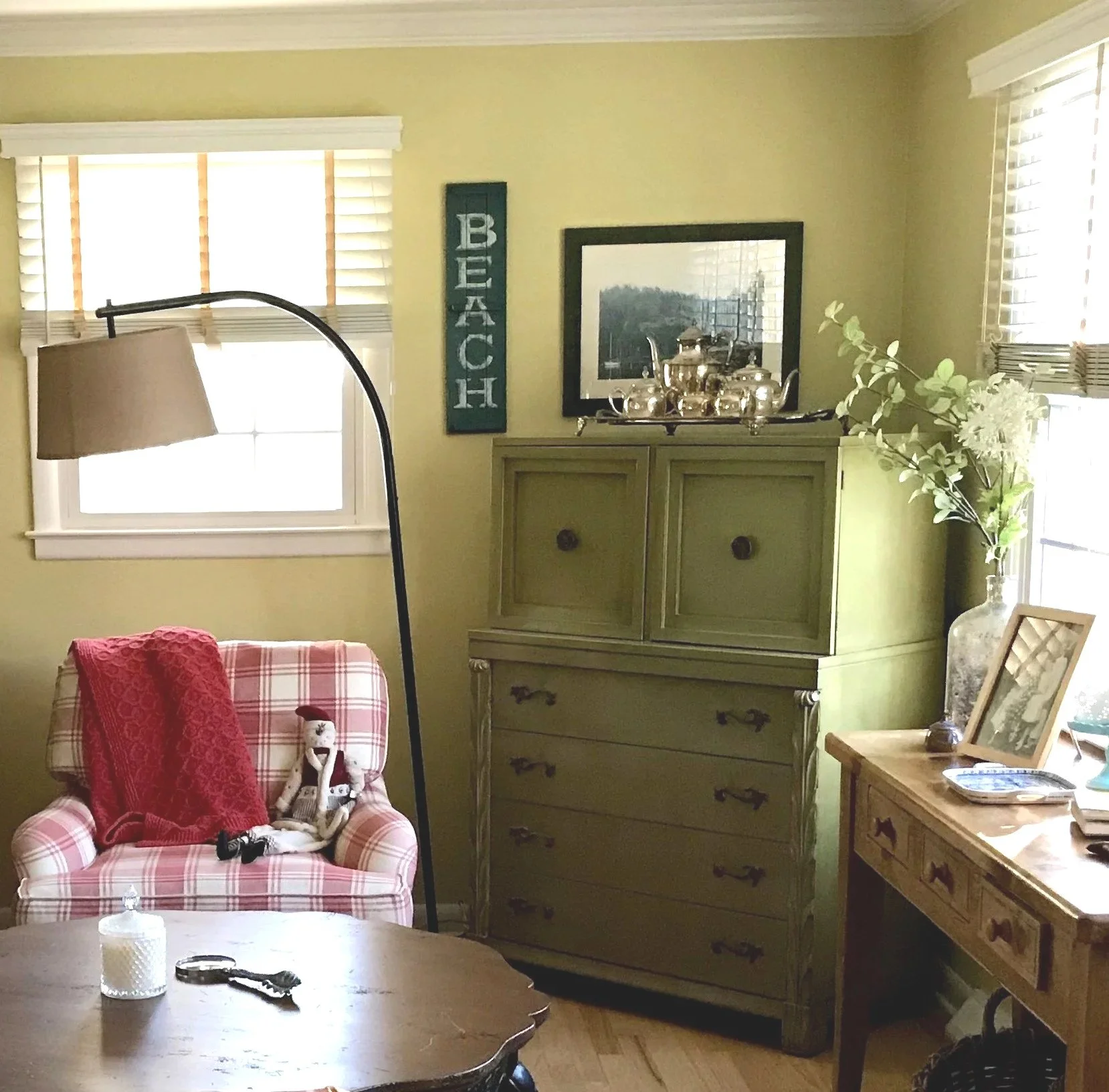 A red and white plaid armchair with a red knit throw beside an arched floor lamp, next to a green dresser topped with a silver tea set