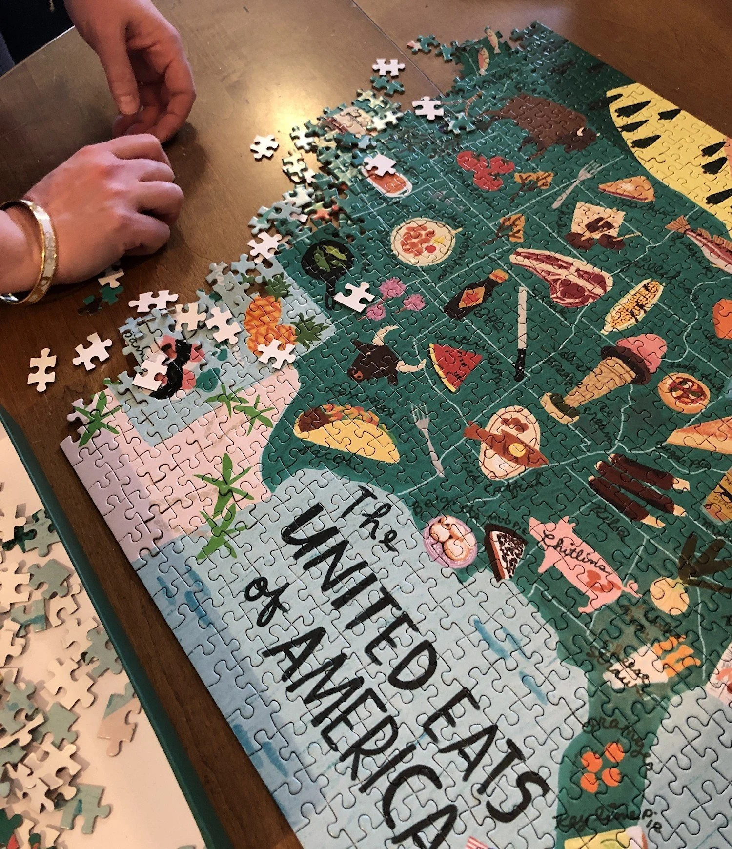 Hands sorting loose pieces beside a nearly finished jigsaw puzzle map of the United States, illustrated with foods and titled The United Eats of America.