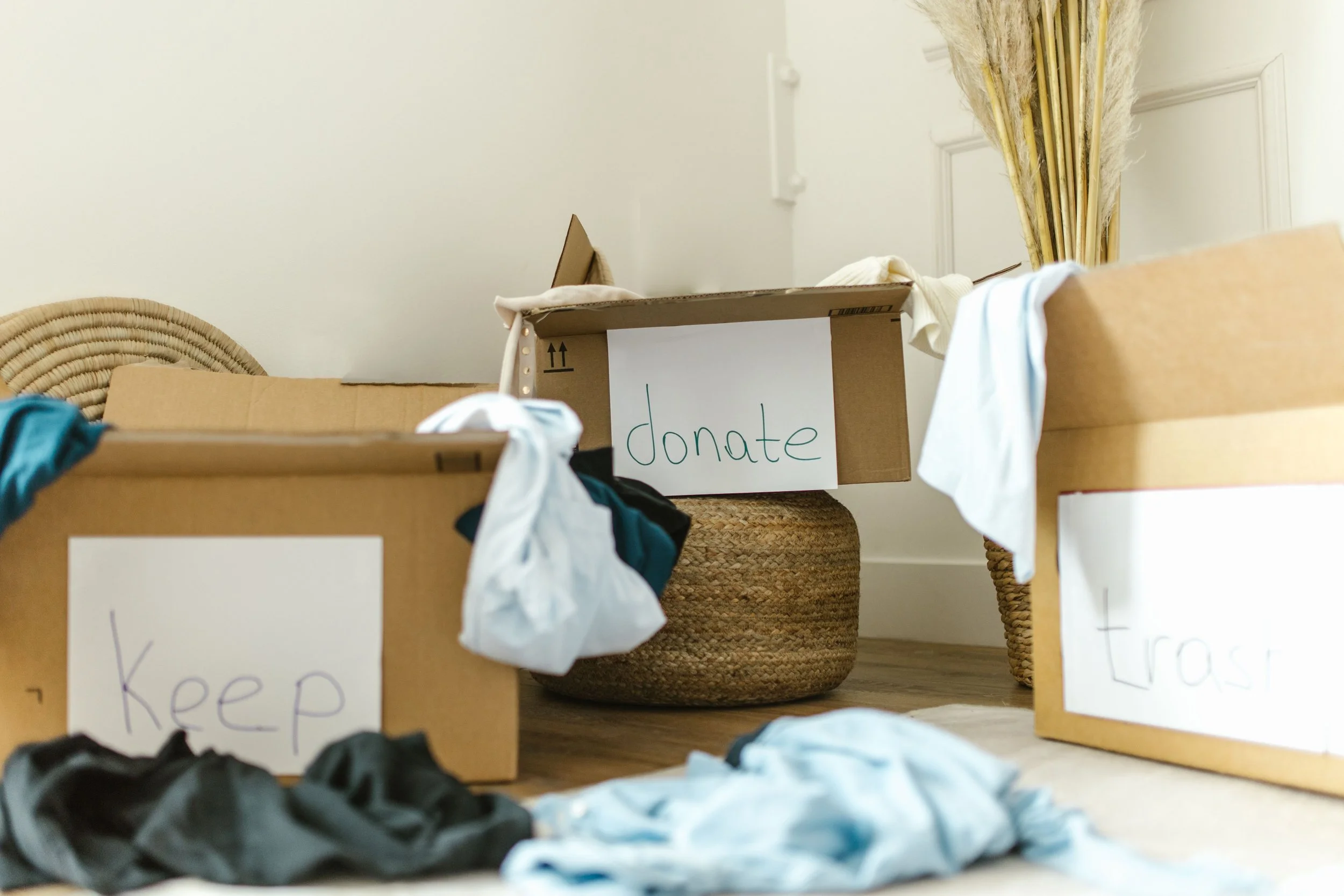 Cardboard boxes labeled keep, donate, and trash with clothing being sorted on the floor while preparing a home for a move