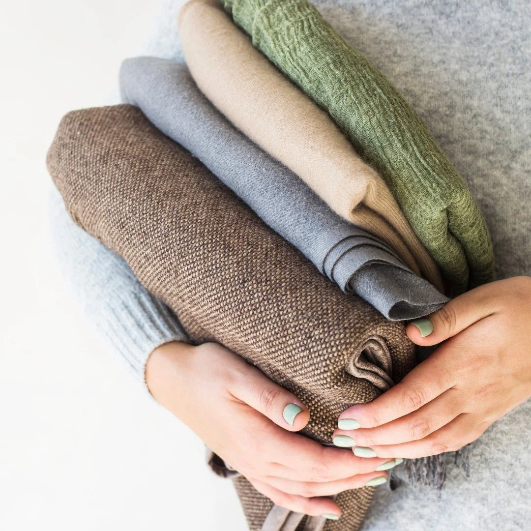 Hands holding a stack of soft folded sweaters in brown, gray, tan, and green