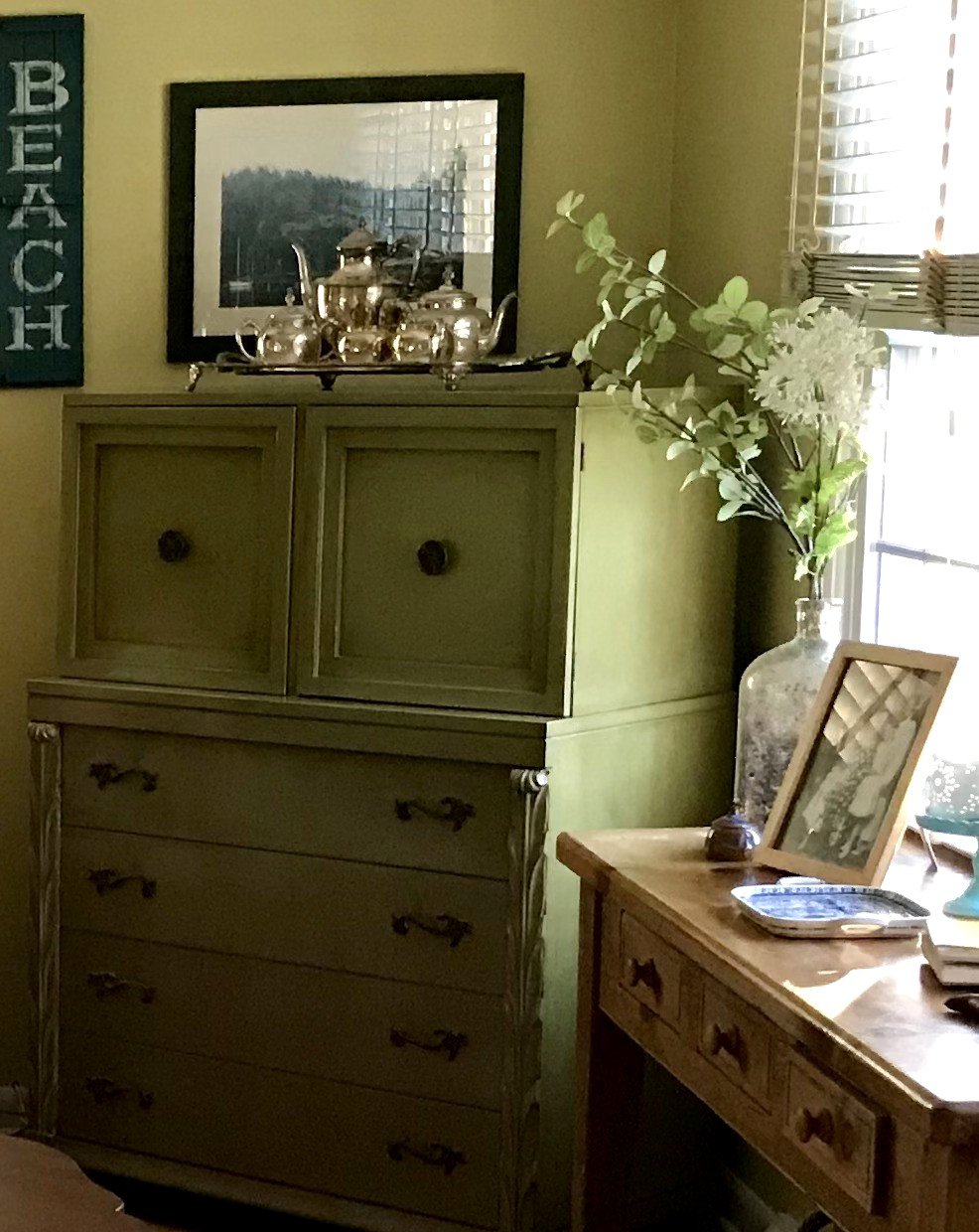 A silver tea set on a green painted dresser beside a vase of white flowers and a vintage framed family photo on a wooden desk