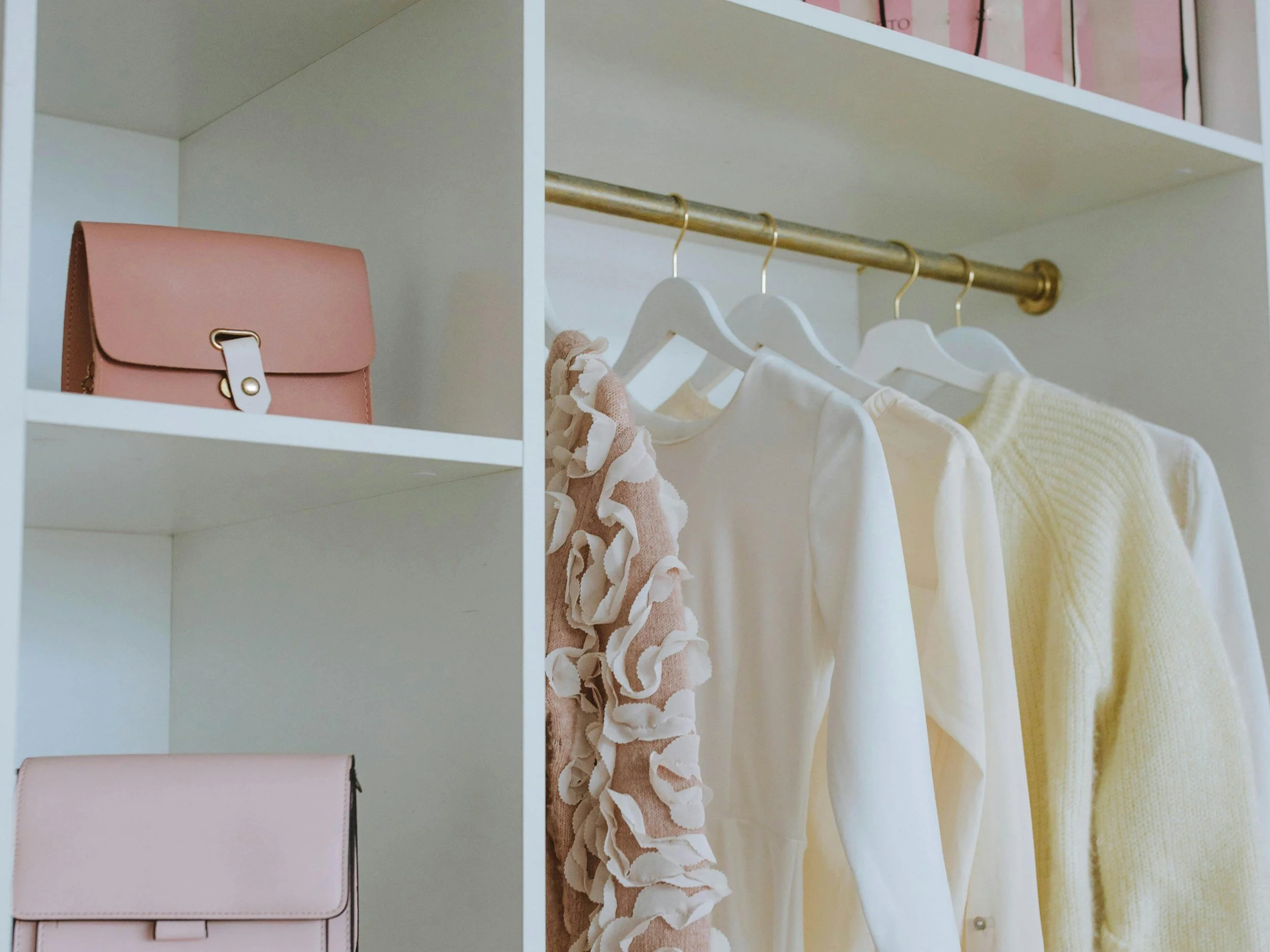 A bright white closet with a cream knit sweater and blouses on gold hangers beside open shelves holding leather handbags