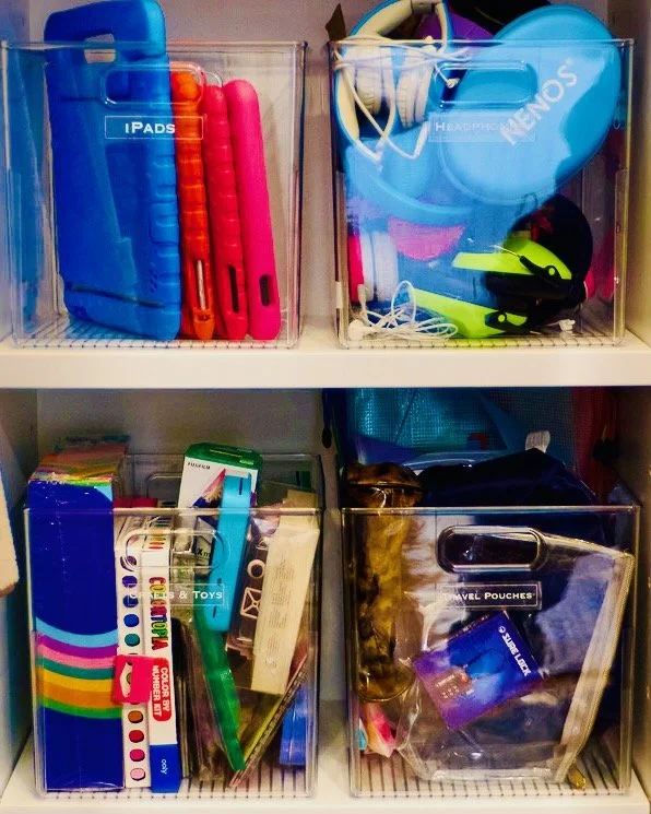 Clear open bins labeled iPads, Headphones, Crafts and Toys, and Travel Pouches on white closet shelves