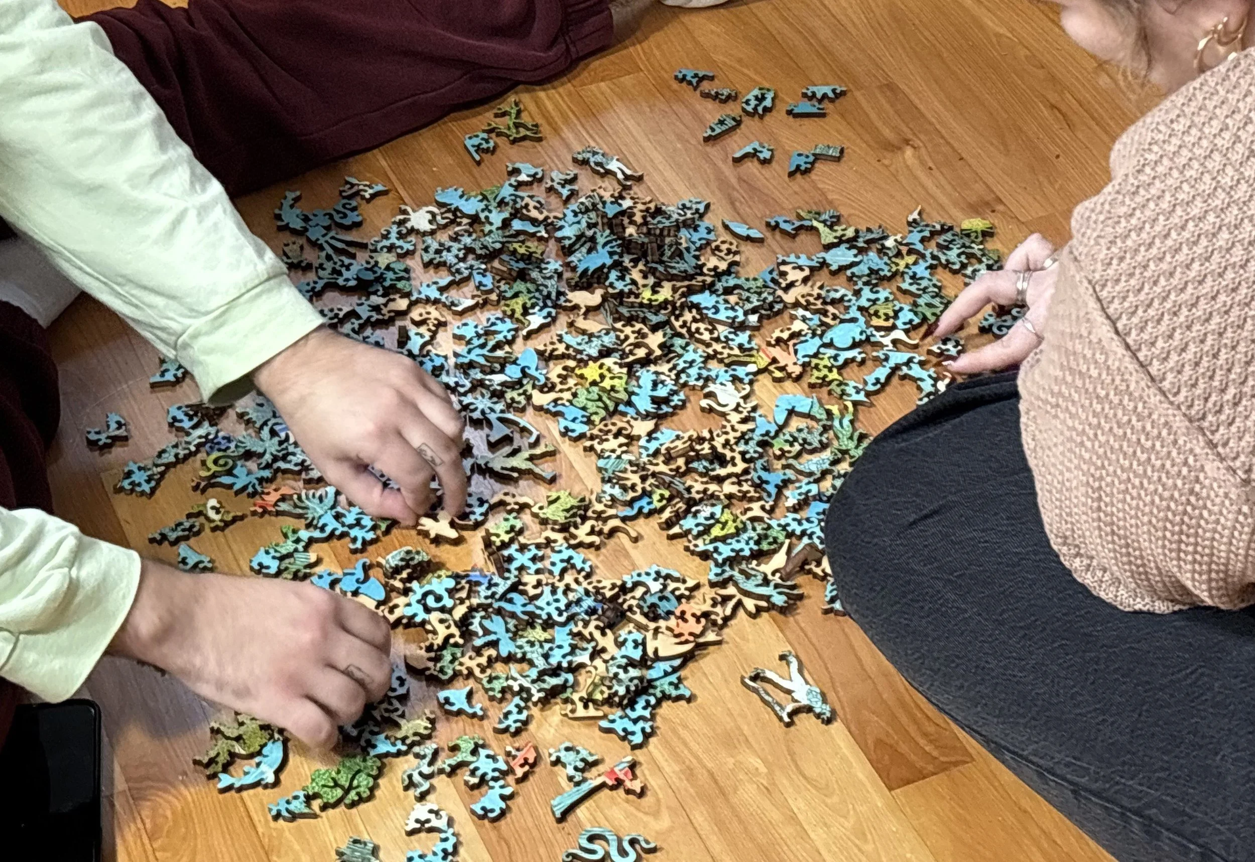 Two people sitting on a wood floor sorting through a large pile of wooden puzzle pieces