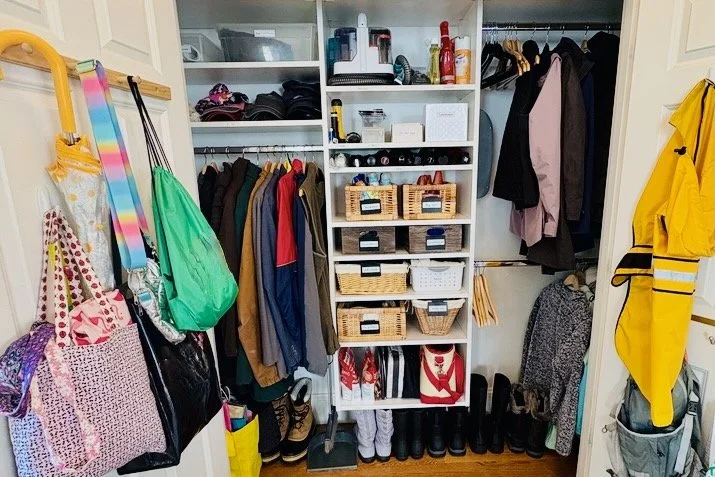 Family coat closet with bags on door hooks, labeled open baskets on shelves, and boots lined up on the floor