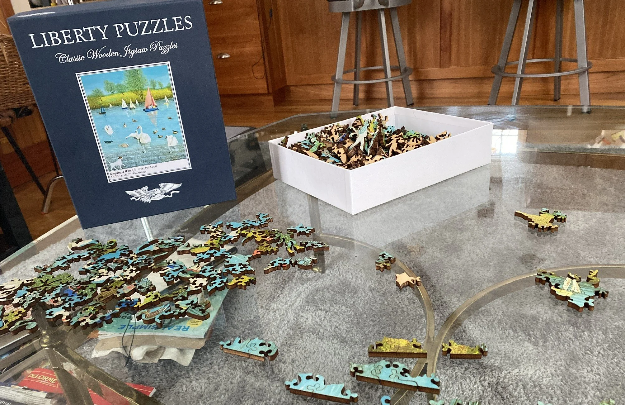 A wooden jigsaw puzzle in progress on a glass coffee table, with the box top standing up and a tray of unsorted pieces