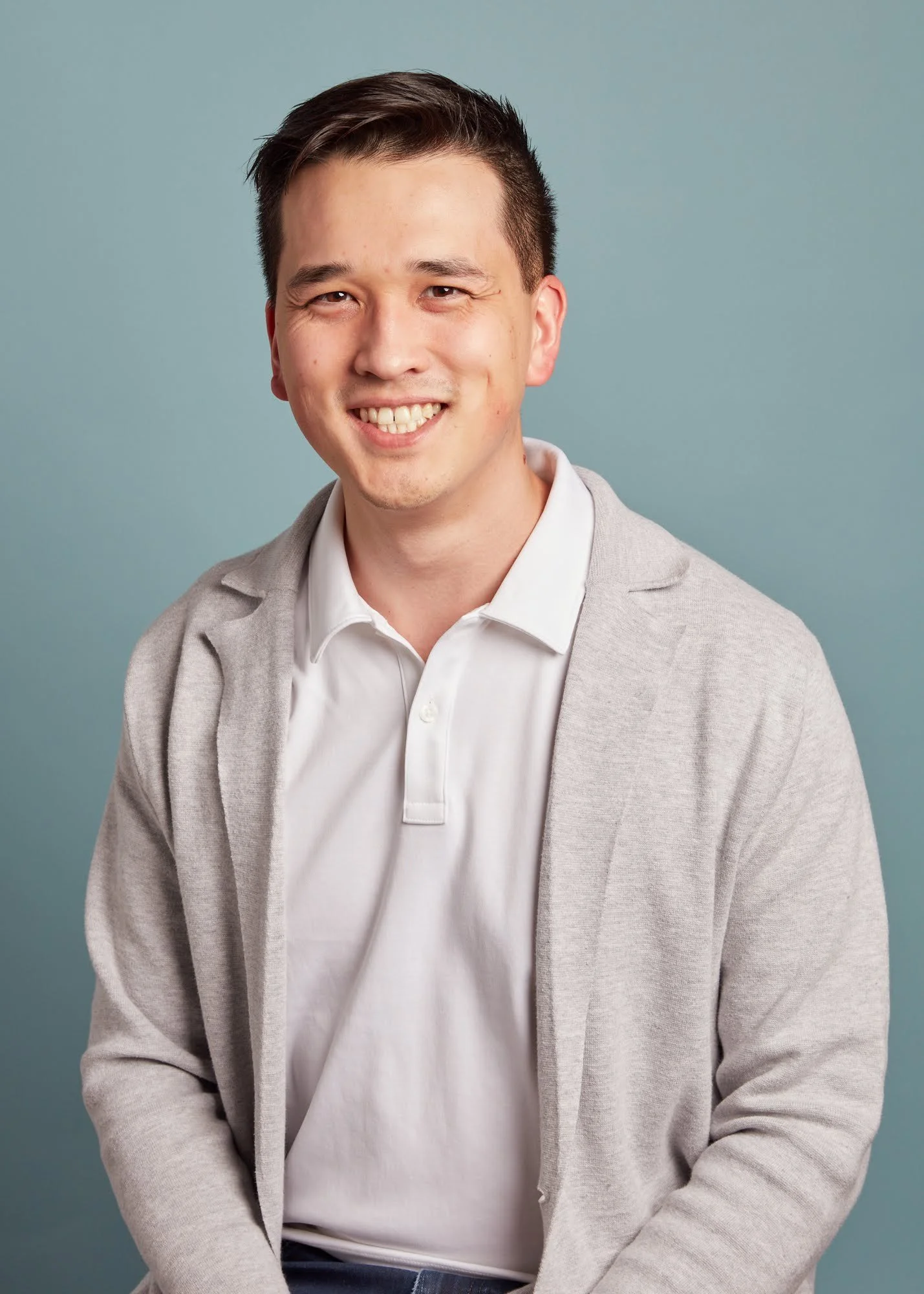 Portrait of a young man with dark hair and a friendly smile, wearing a white polo shirt and light gray blazer, against a light blue background.