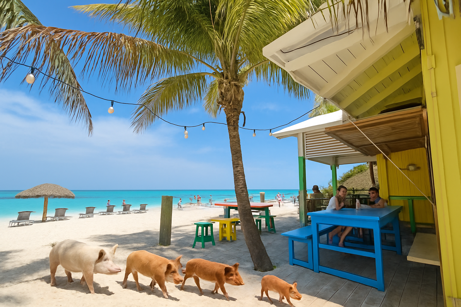 Beach scene with turquoise water, sand, and lounge chairs, shaded by a thatched umbrella. A palm tree is in the foreground with string lights hanging overhead. There are people in the distance and two people sitting at a blue table under a shaded structure, with colorful stools nearby.