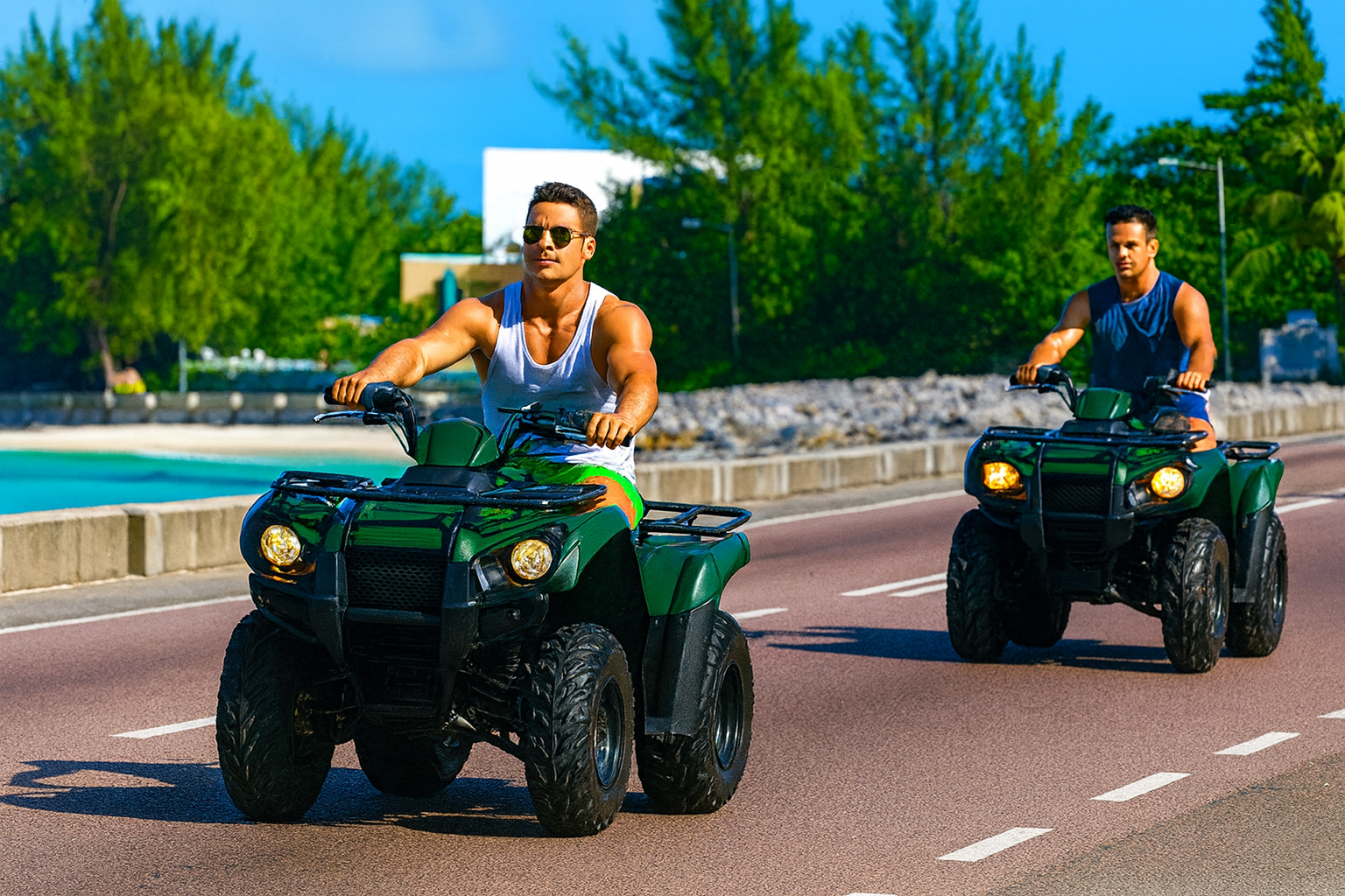 Two women riding all-terrain vehicles (ATVs) on a paved road outdoors on a sunny day.