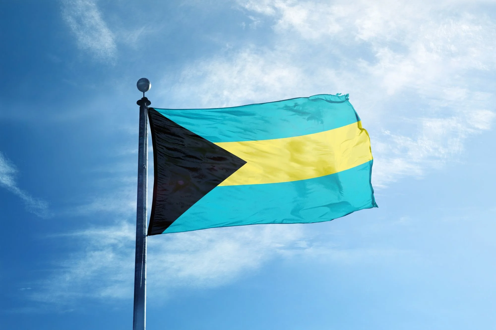 Flag of the Bahamas with a light blue, yellow, and black design, flying against a partly cloudy sky.