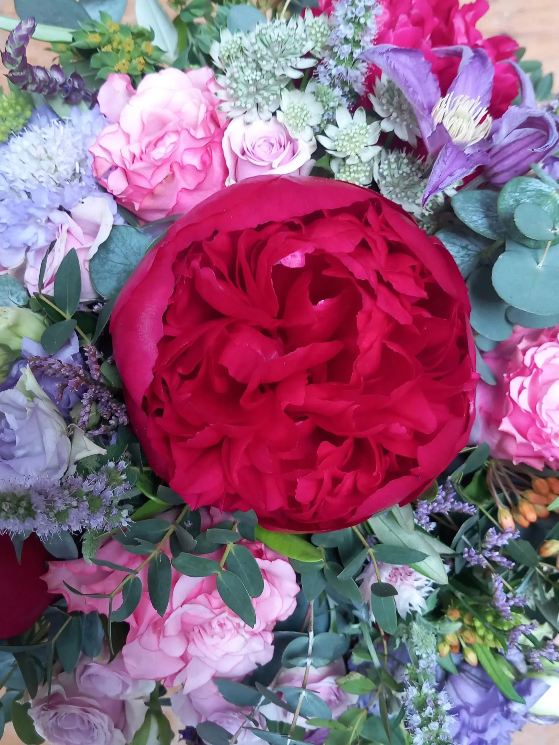 A colorful mixed wedding bouquet of flowers including a large red peony, pink roses, purple clematis and vgreen foliage.
