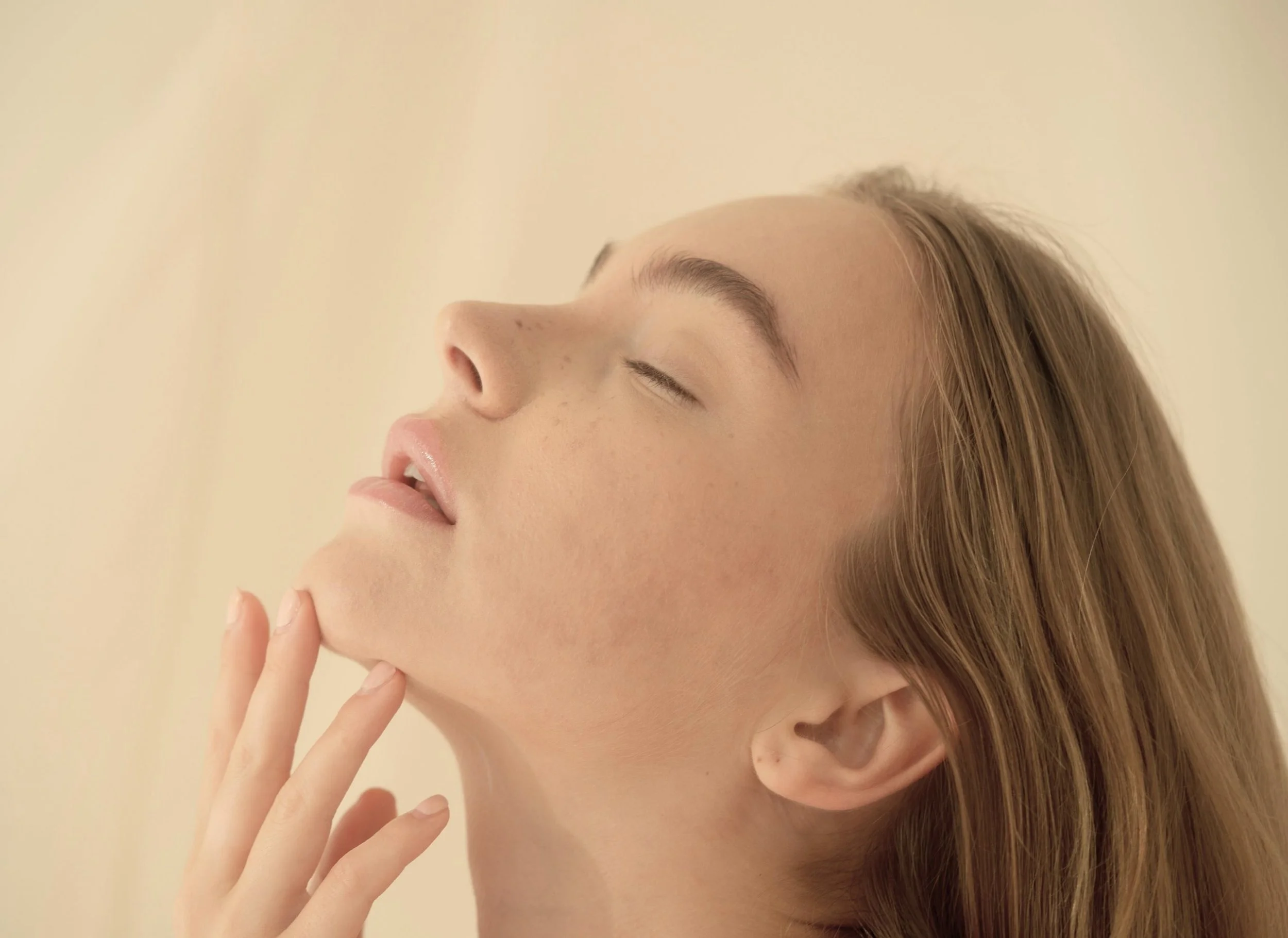 Close-up of a woman with fair skin and light brown hair with her eyes closed, gently touching her chin with her fingers, against a neutral background.