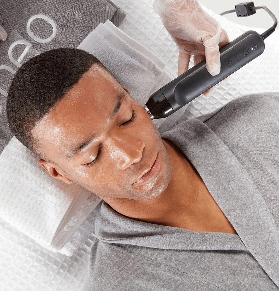 A man lying down with eyes closed, receiving a facial treatment with a black handheld device.