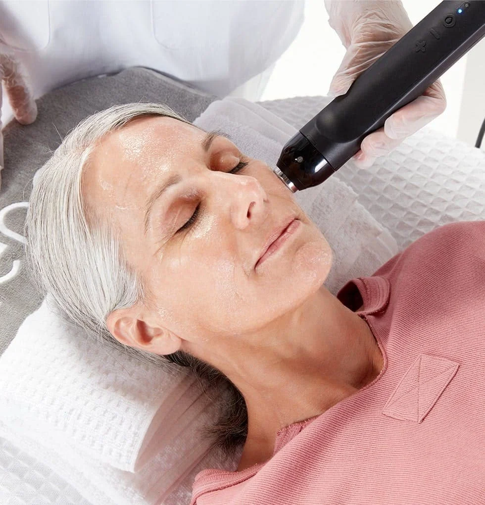 A woman with gray hair relaxing with her eyes closed while a professional administers a facial treatment using a handheld device.
