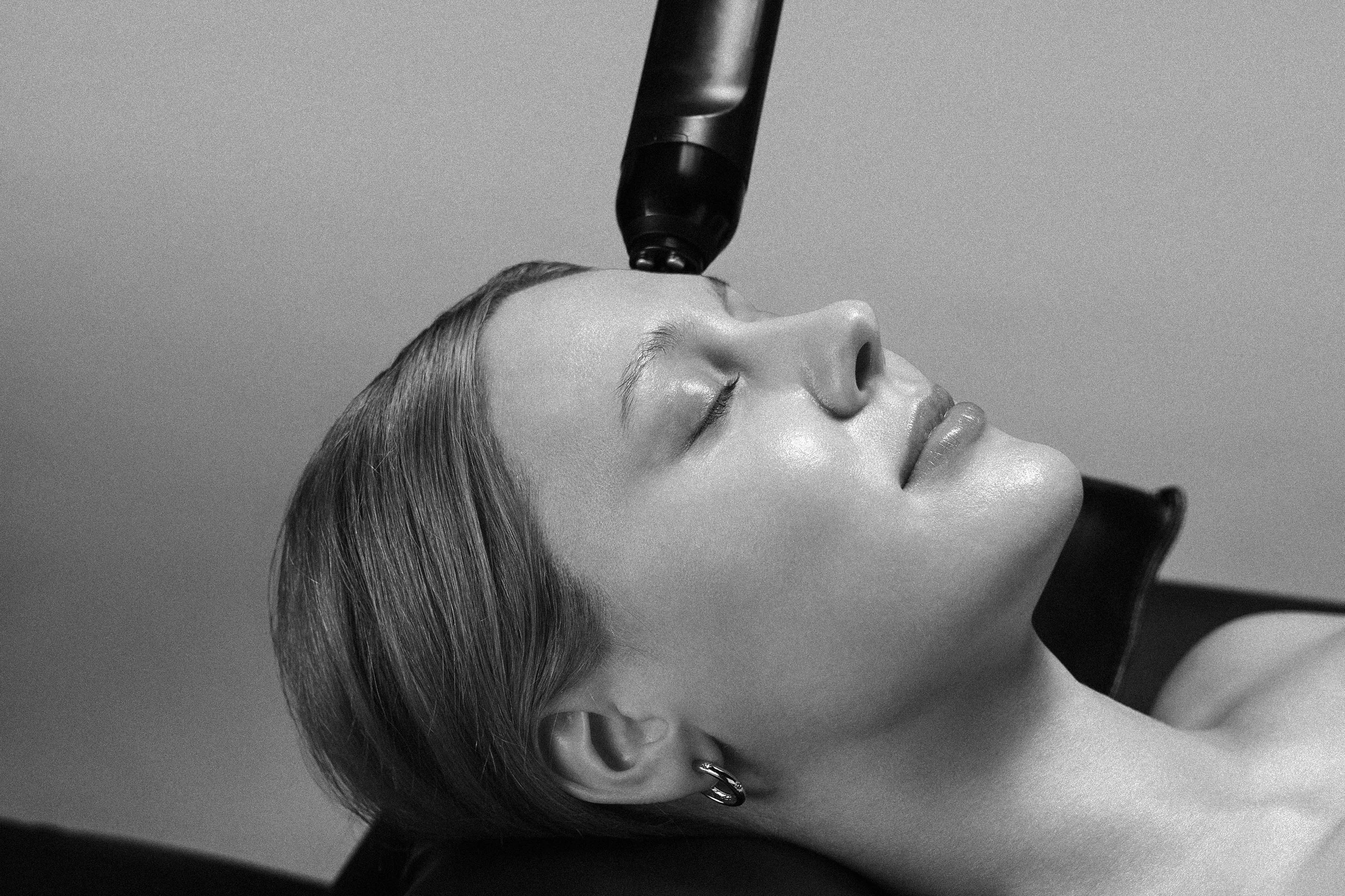 A woman lying down with her eyes closed while receiving a cosmetic or skincare treatment from a professional holding a device above her face.