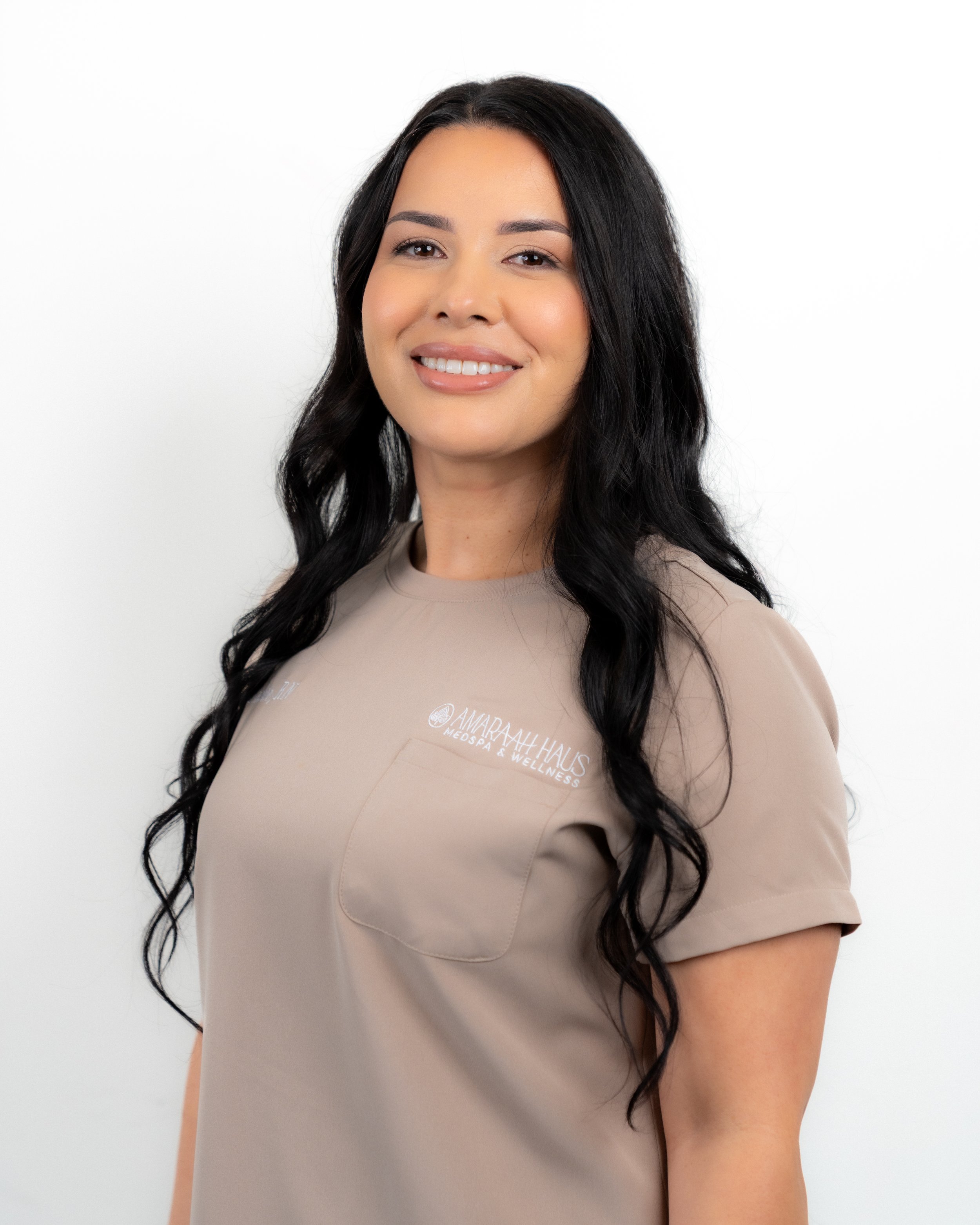 A woman with long dark hair smiling, wearing a beige top with the logo 'Amara Nah Haus Medspa & Wellness' on the chest, standing against a white background.