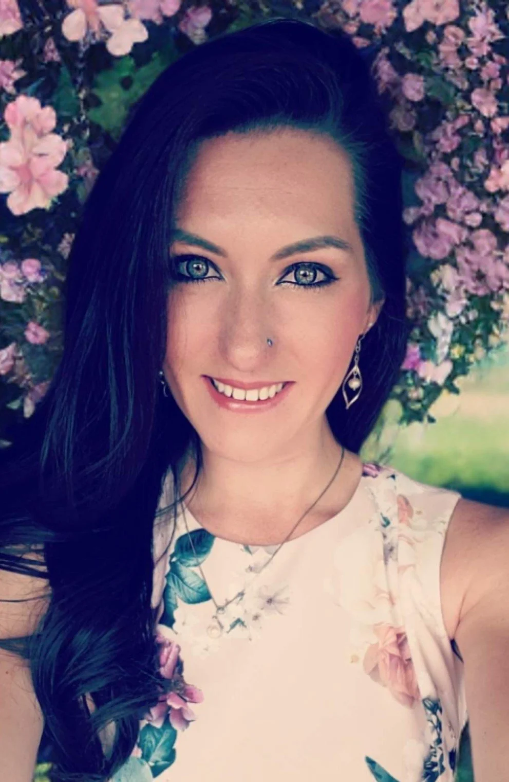 A woman with long dark hair, blue eyes, and a nose piercing, smiling and taking a selfie in front of pink flowers.