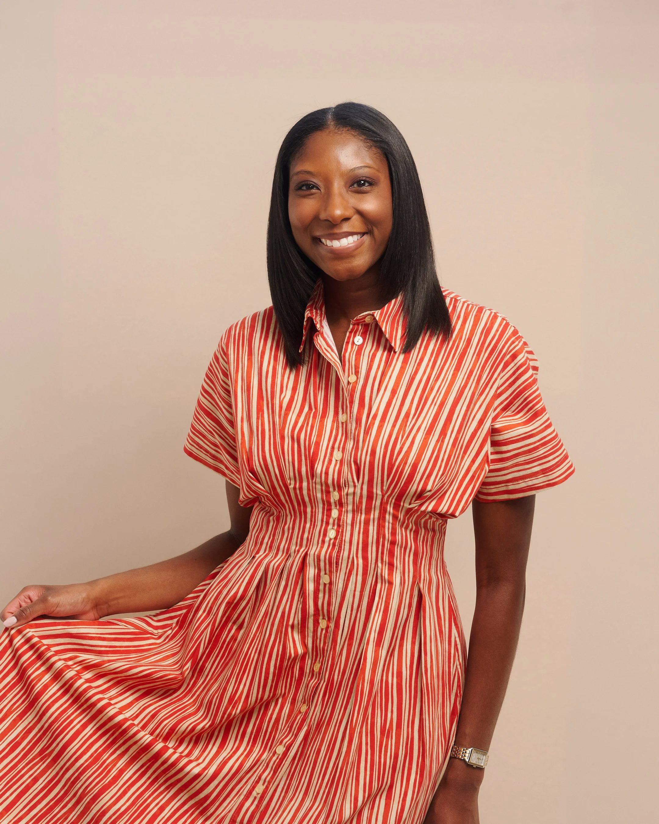 Portrait of Allison— A Black woman with straight black hair wearing a striped red and beige dress, smiling and sitting against a plain beige wall.