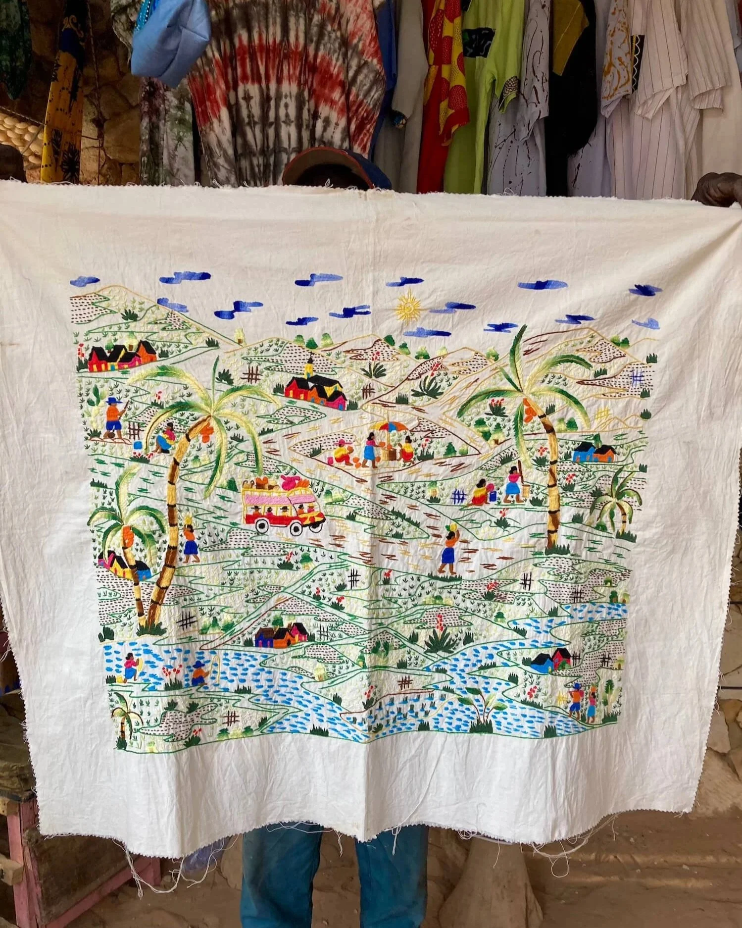 Tissu brodé représentant un paysage rural africain avec des maisons, des palmiers, des figures humaines, des rivières, et des arbres, dans un style coloré et détaillé.