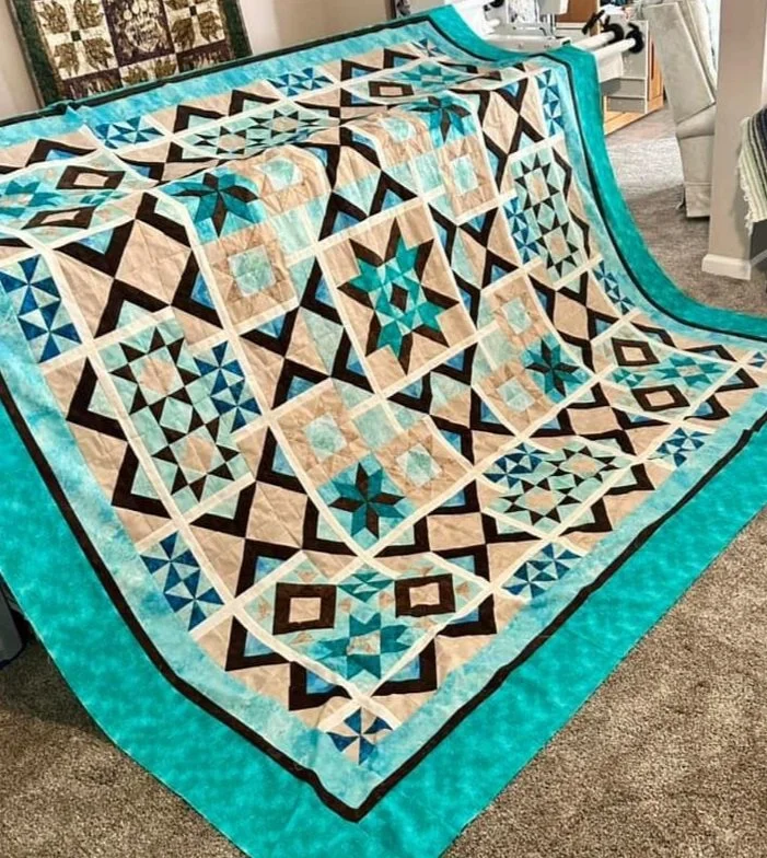 A quilt with a geometric star pattern in shades of beige, blue, and black, bordered by a bright turquoise edge, displayed on a bed or table indoors.