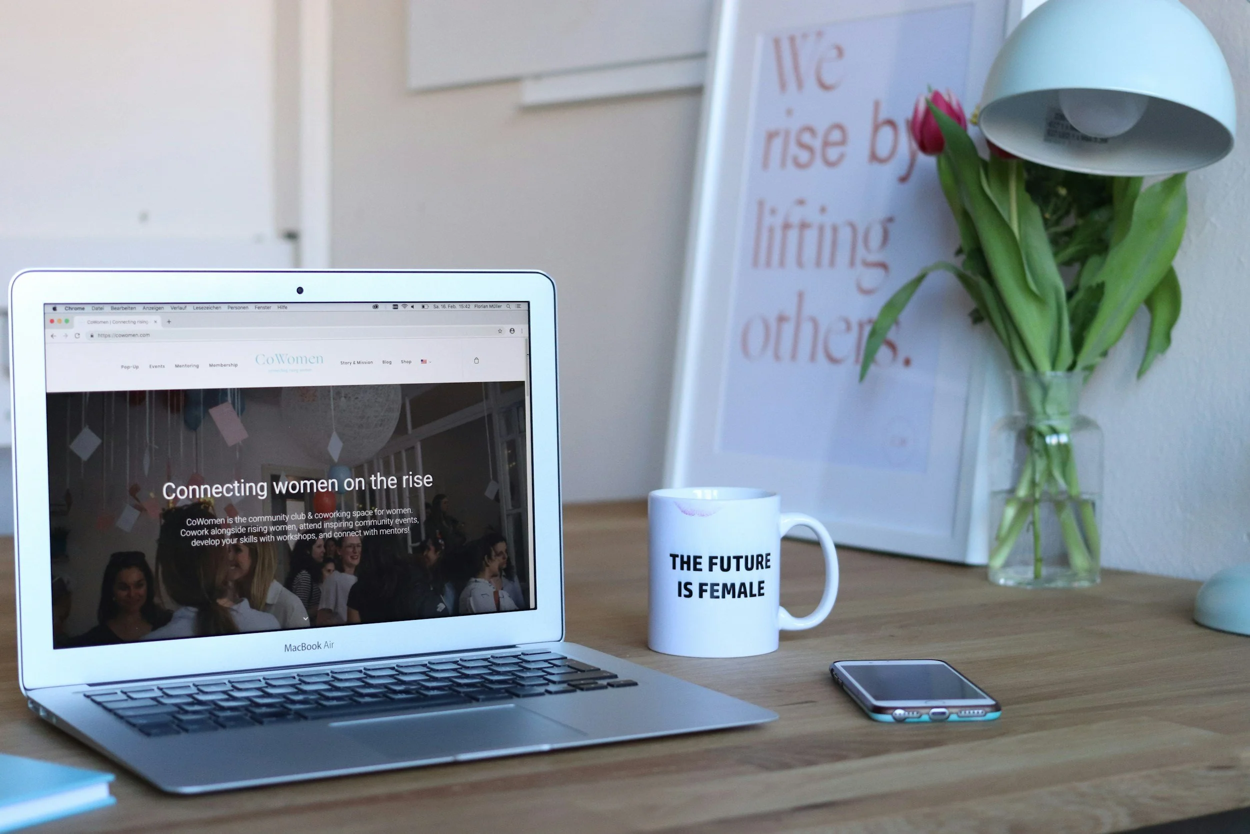 A workspace with a MacBook Air laptop displaying the CoWomen website, a white mug with the text "THE FUTURE IS FEMALE," a smartphone, a glass vase with pink tulips, a framed quote reading "We rise by lifting others," and a desk lamp. The setting appears to be a bright, organized office or home office.