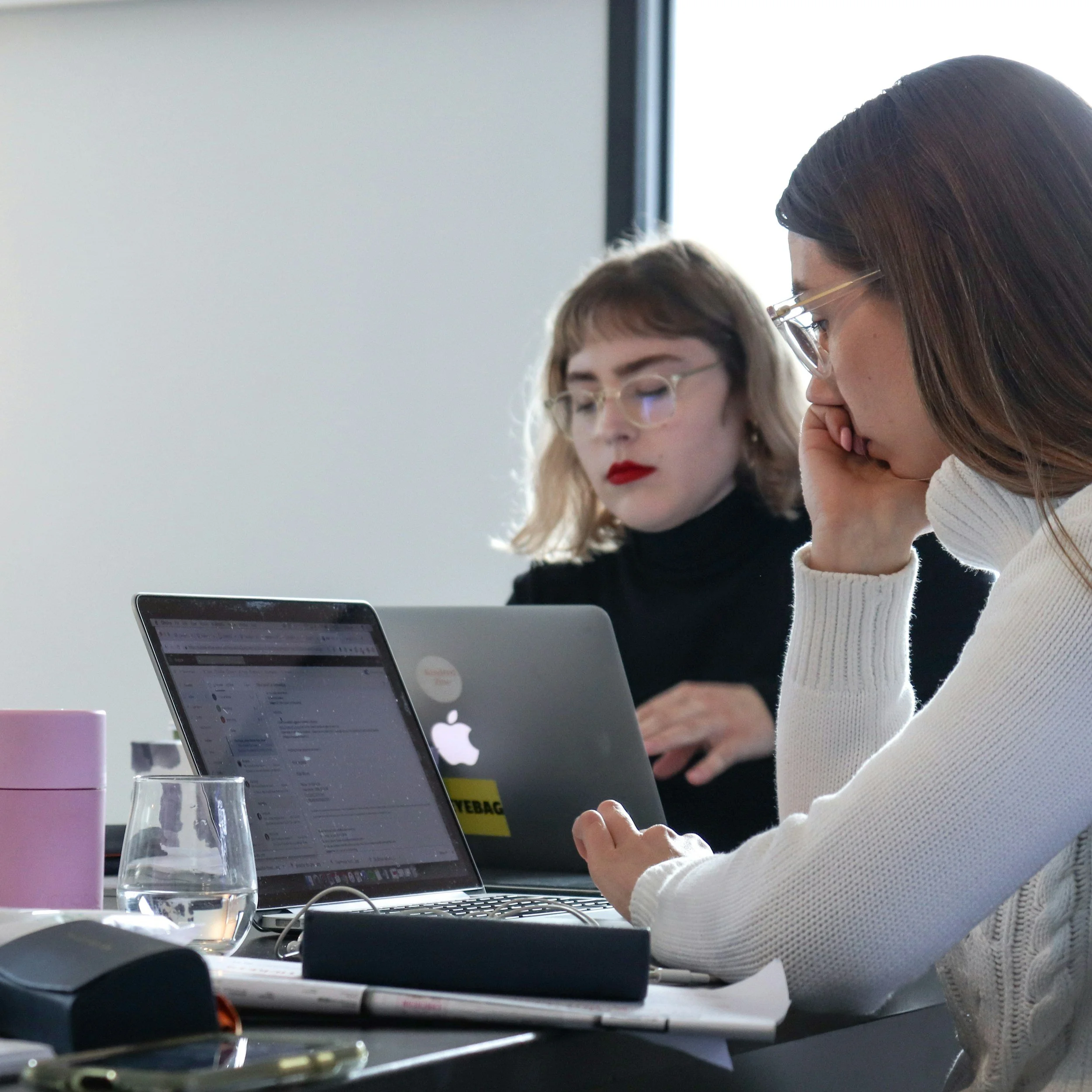 Two women working at a desk with laptops, one in the foreground looking at her screen with her hand on her face, and the other in the background focused on her laptop.