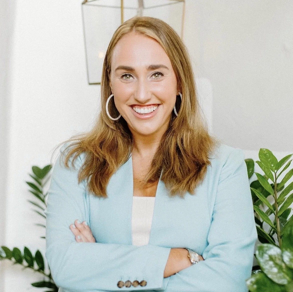 A smiling woman with light brown hair, wearing a light blue blazer and hoop earrings, standing in an office with green plants.