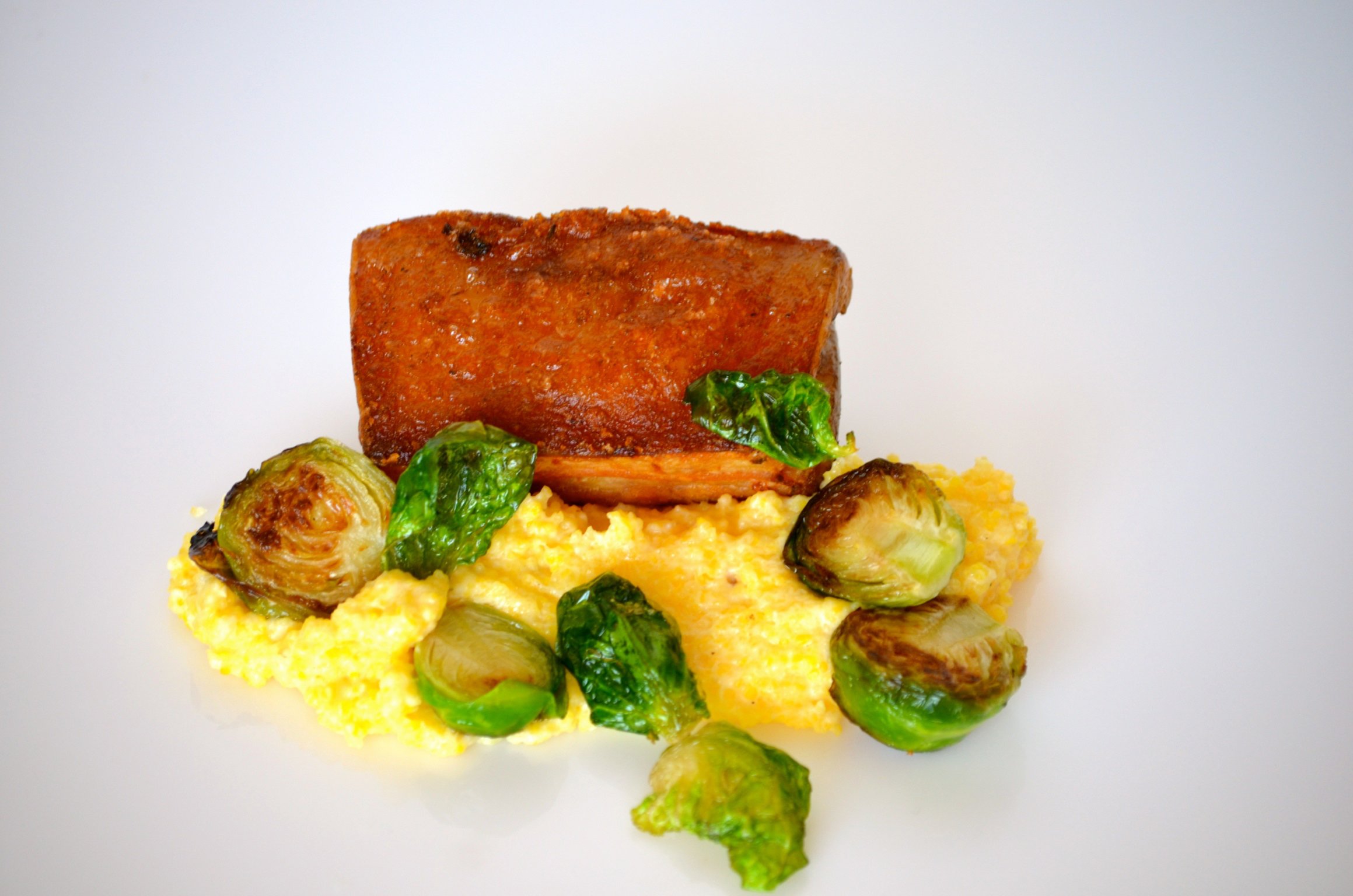A plated dish featuring a rectangular piece of cooked salmon, garnished with green herbs, served over a bed of yellow mashed potatoes with green Brussels sprouts around it.