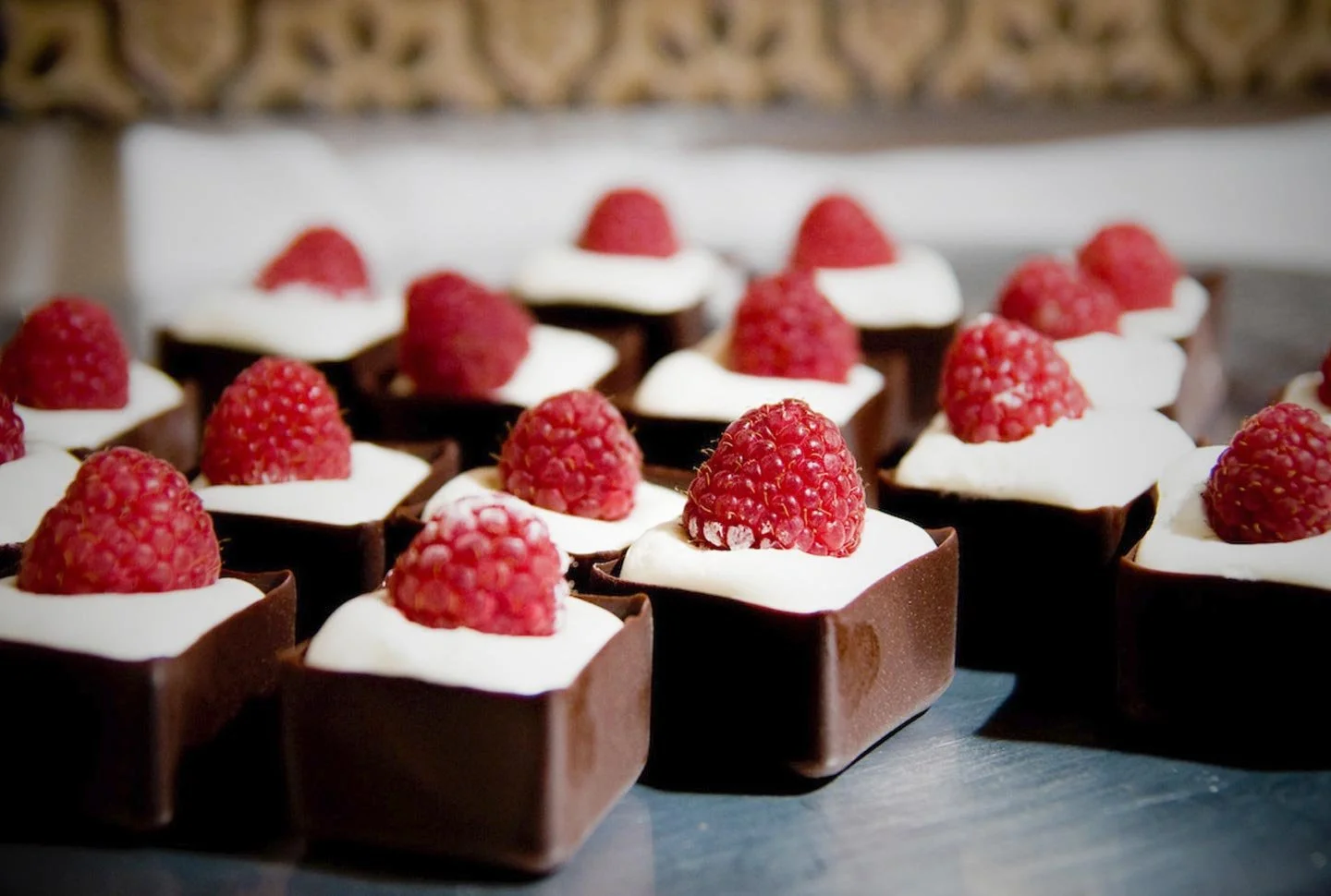 Square chocolate cups topped with white frosting and a raspberry on each.