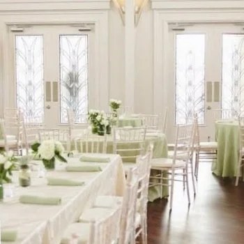 Elegant dining room with round tables dressed in white tablecloths and light green accents, surrounded by white chairs, large windows with sheer curtains, and minimalistic decor.