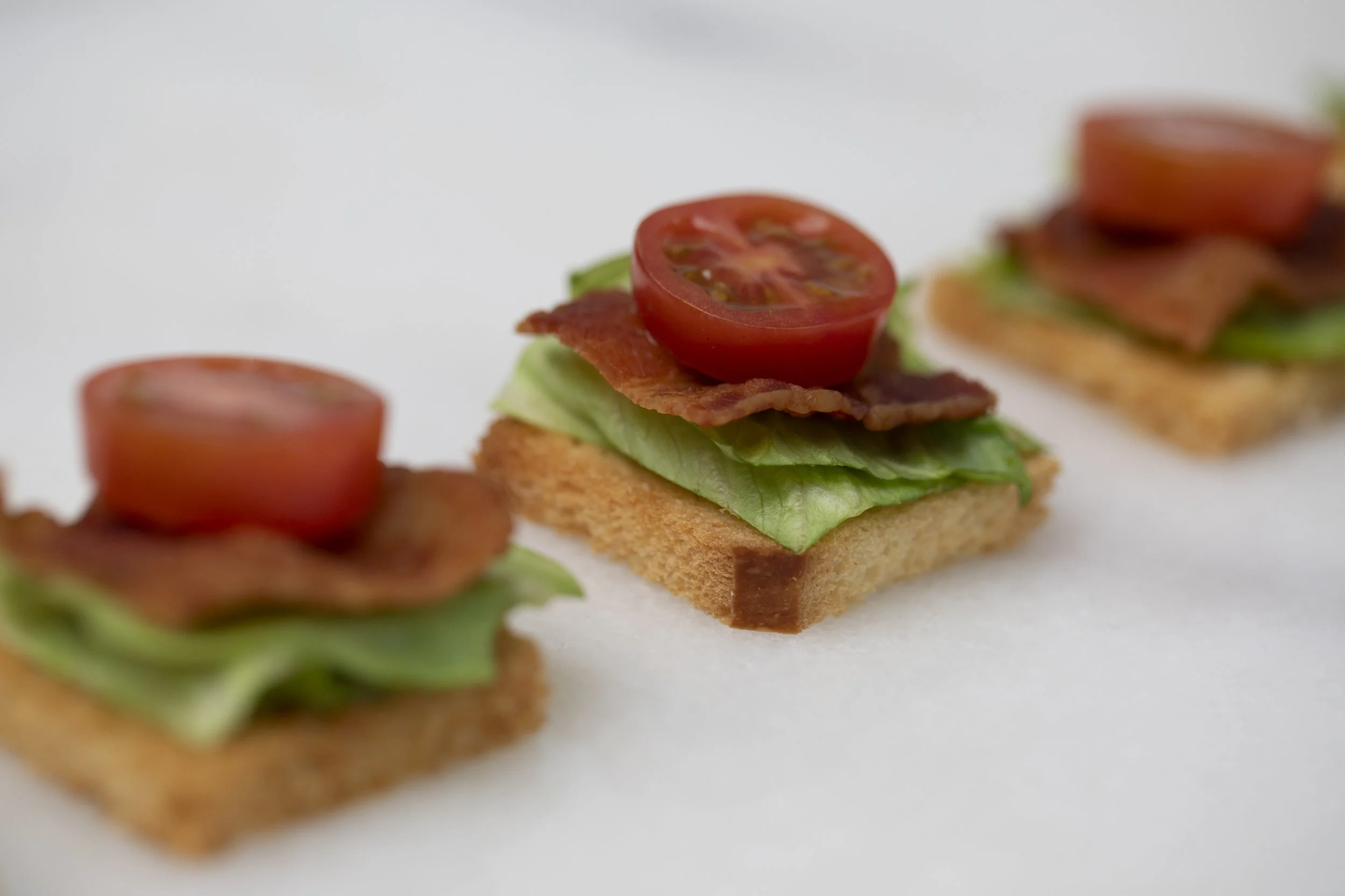Close-up of a small appetizer sandwich with a toasted bread base, topped with lettuce, bacon, and halved cherry tomatoes, arranged on a white surface.