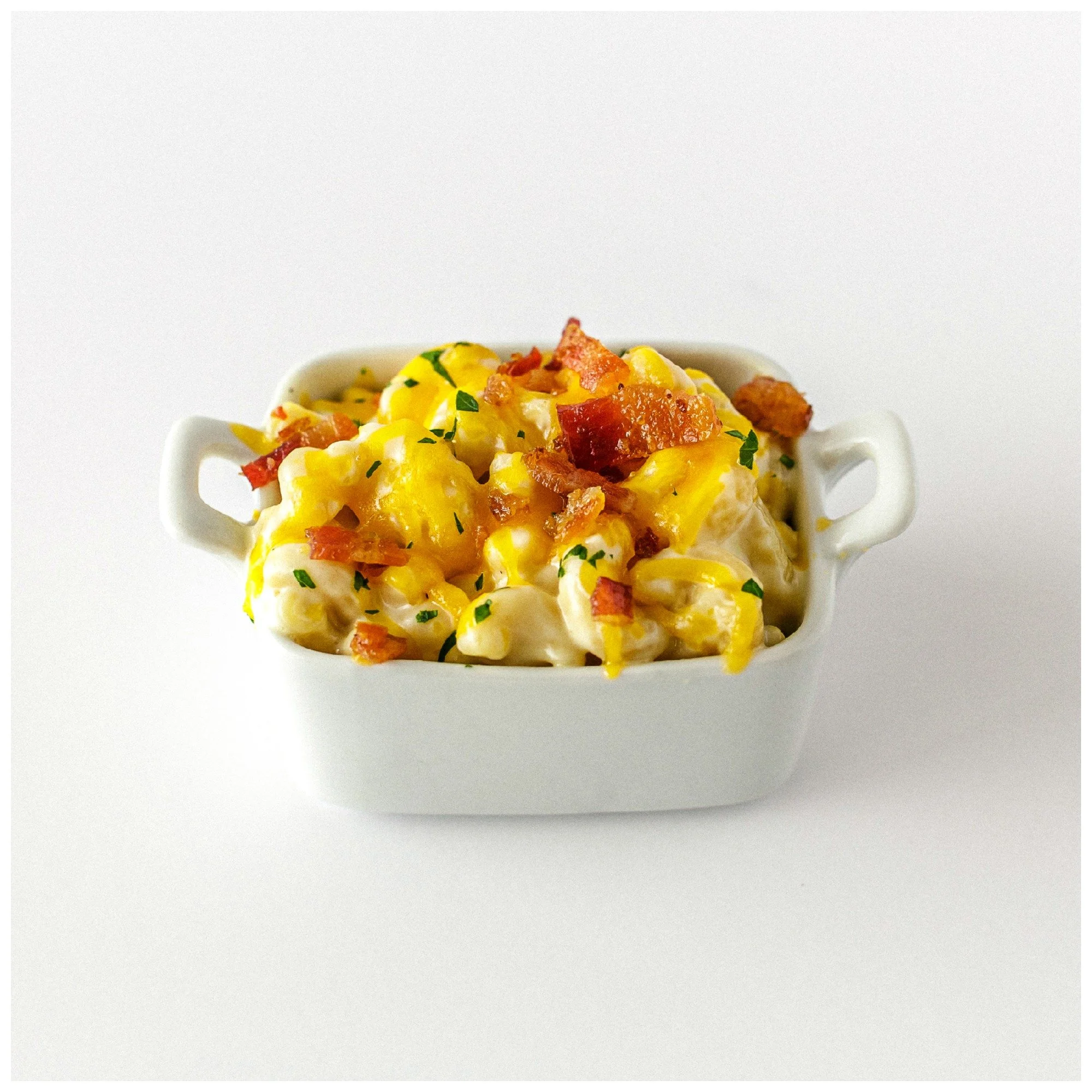 A small white ceramic dish filled with creamy mac and cheese topped with crispy bacon bits and garnished with chopped herbs, set against a plain white background.