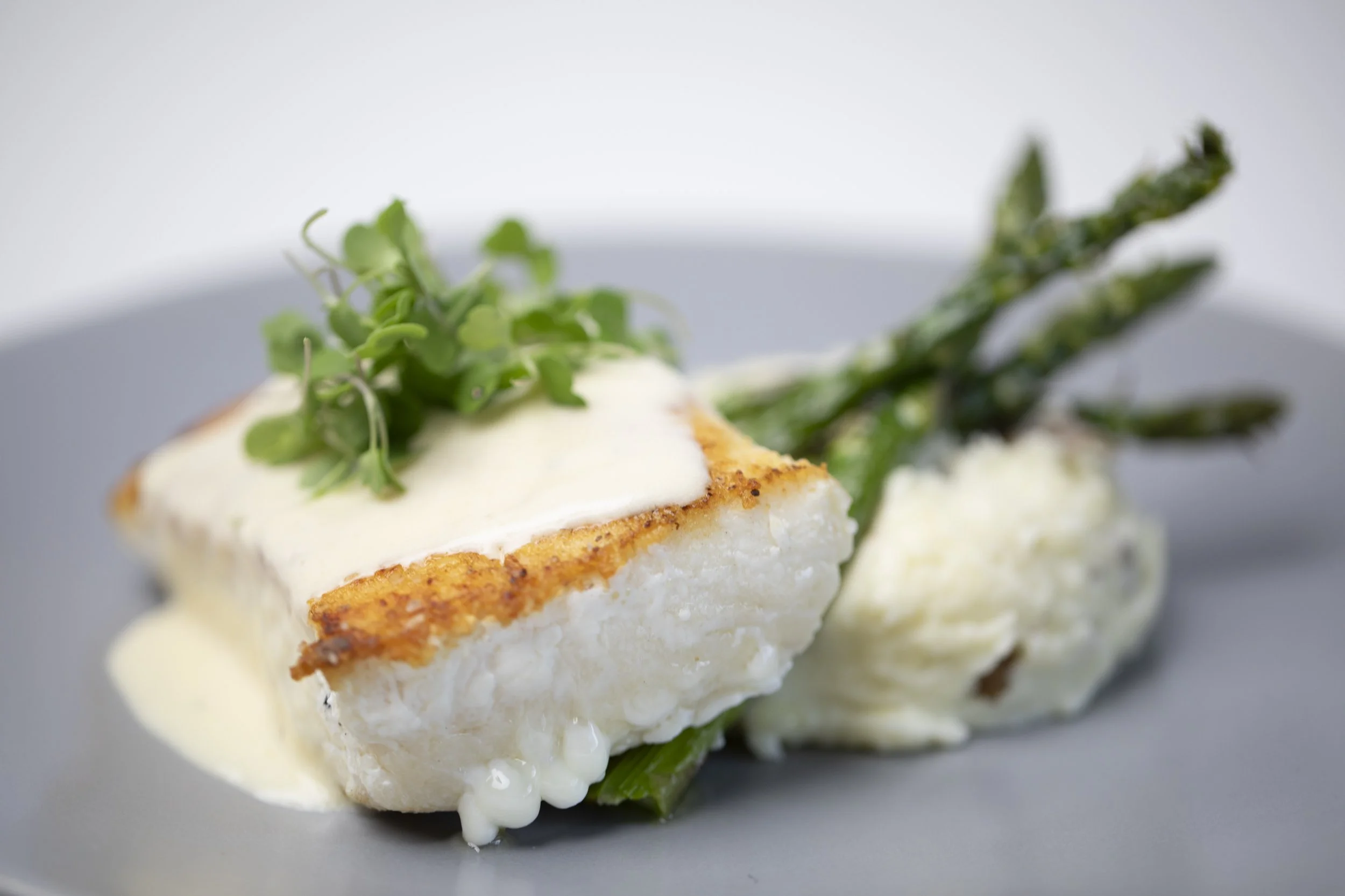 Close-up of a piece of white fish topped with a creamy sauce and garnished with microgreens and green vegetables, served on a gray plate.