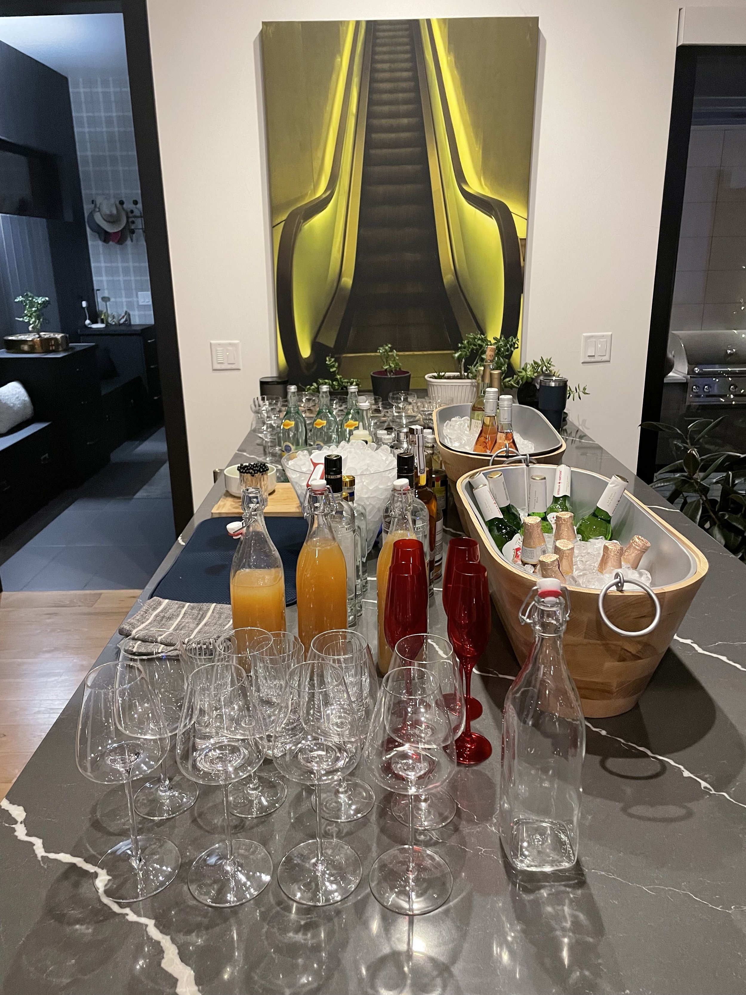 A table set up with bottles of various drinks, glasses, and ice, indicating a gathering or party, with a large colorful escalator mural in the background.