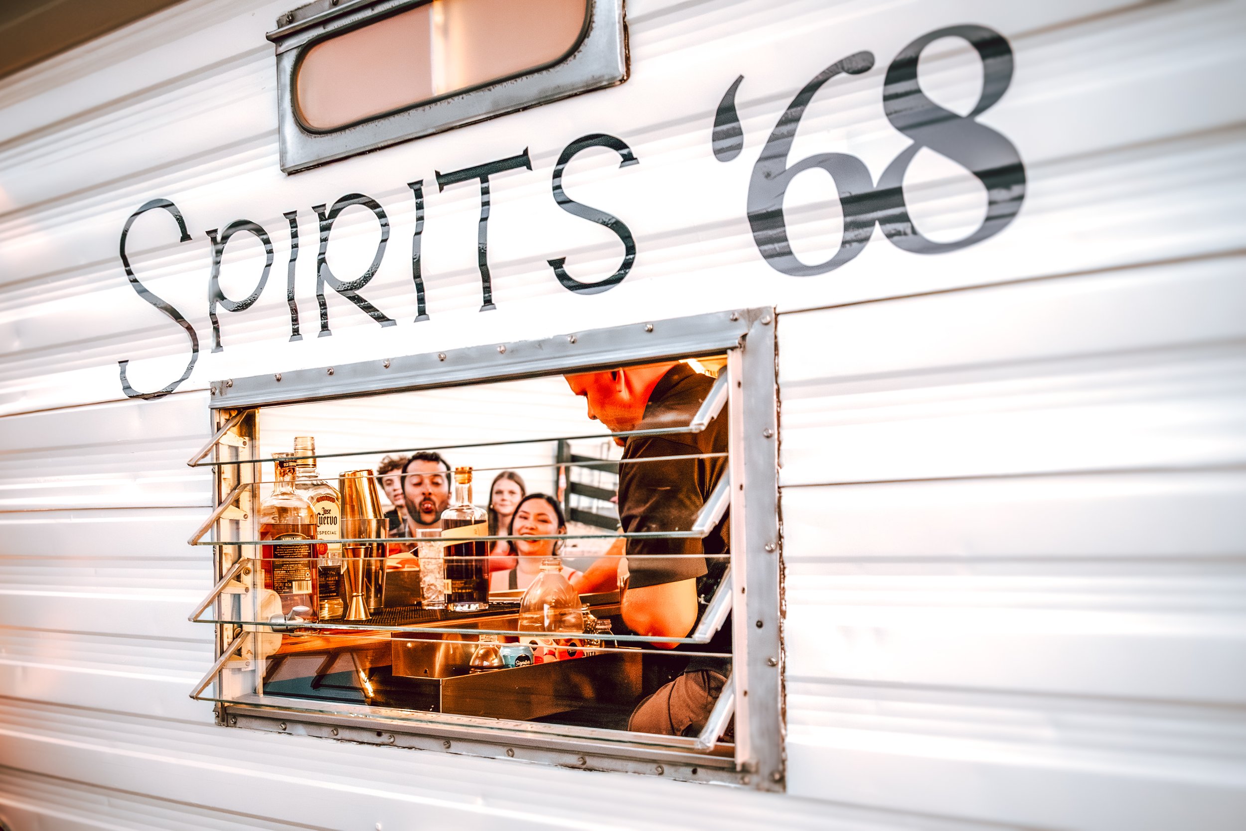 A group of people enjoying drinks at a food truck named "Spirits '68" with a bartender preparing beverages through an open window.