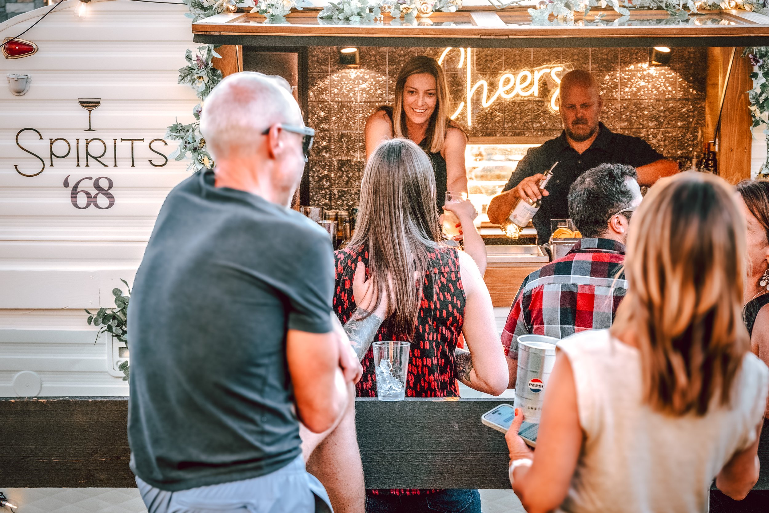 People at an outdoor bar with a bartender pouring drinks and a neon 'cheers' sign, surrounded by floral decorations and signage reading 'SPIRITS '68'.