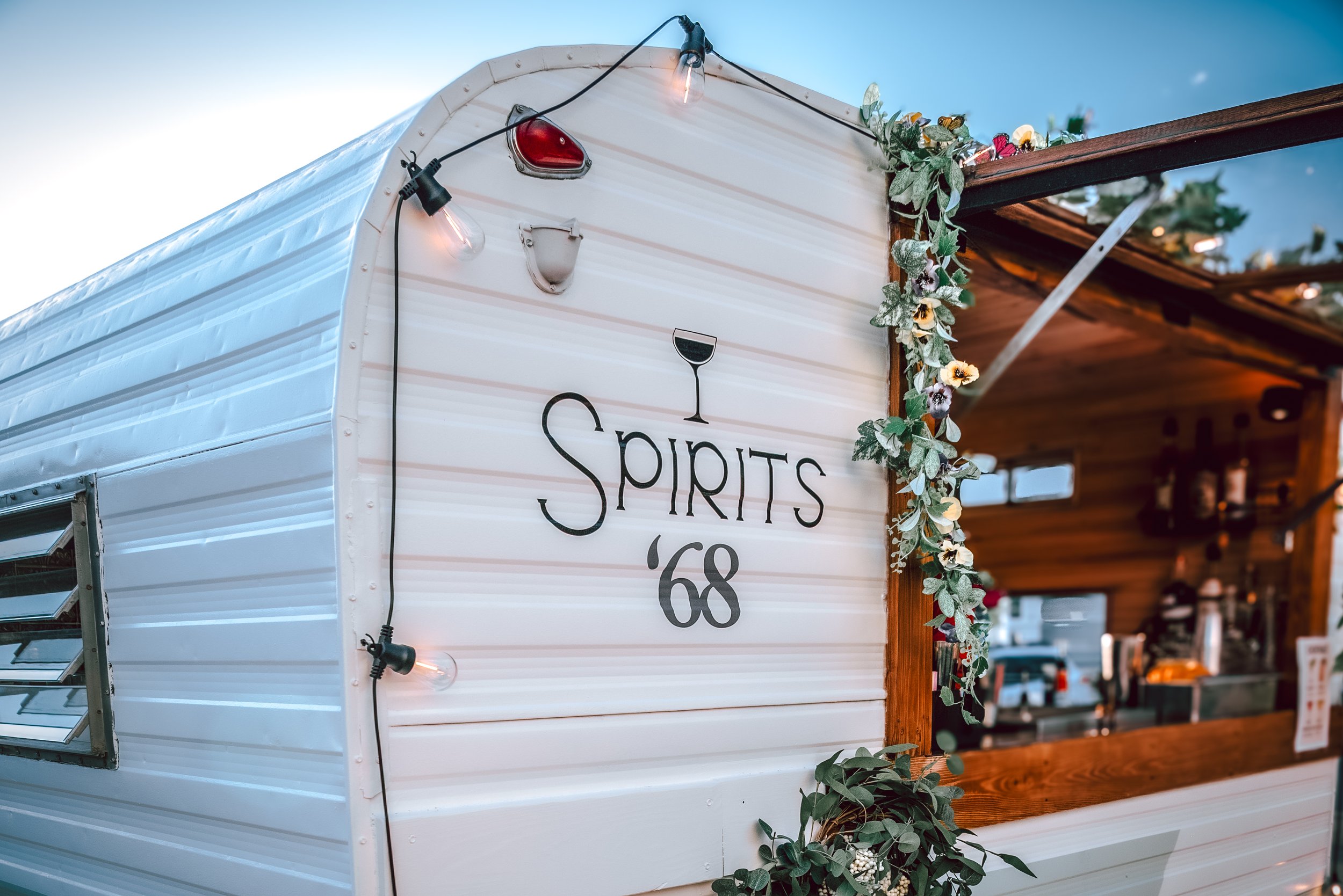 A vintage trailer decorated with string lights and green foliage, with a sign that reads "Spirits '68" featuring a wine glass icon, set against a partly cloudy sky.