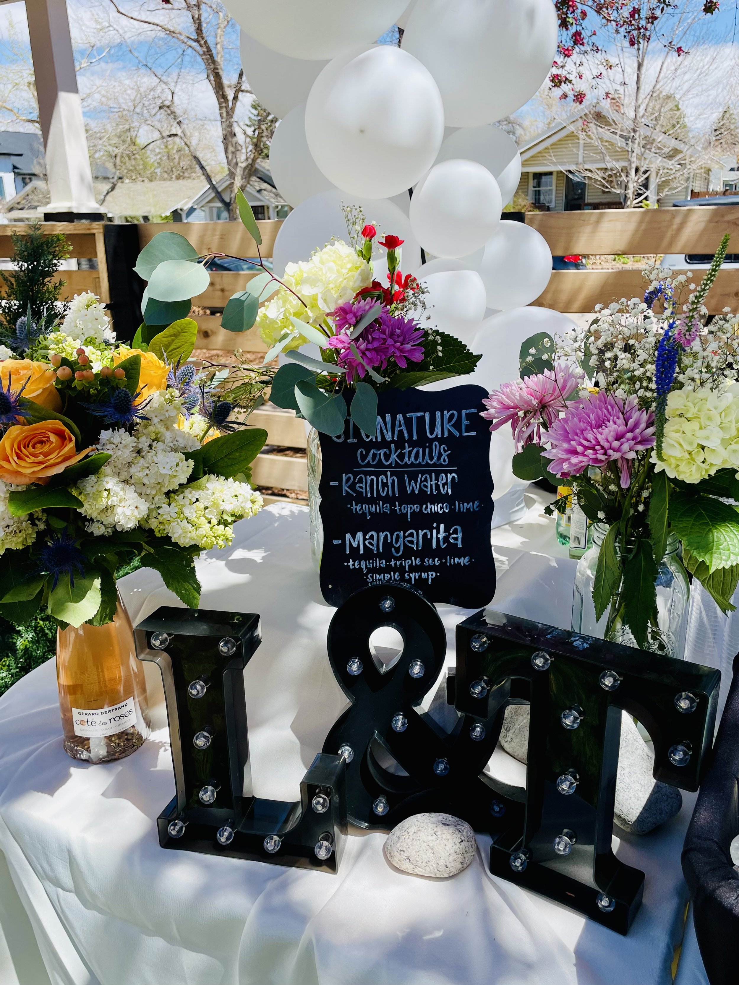 Table decorated with flower arrangements, white balloons, and black marquee letters spelling 'LOVE' with small light bulbs. A chalkboard sign lists signature cocktails: ranch water and margarita. The background shows a clear sky and some trees.