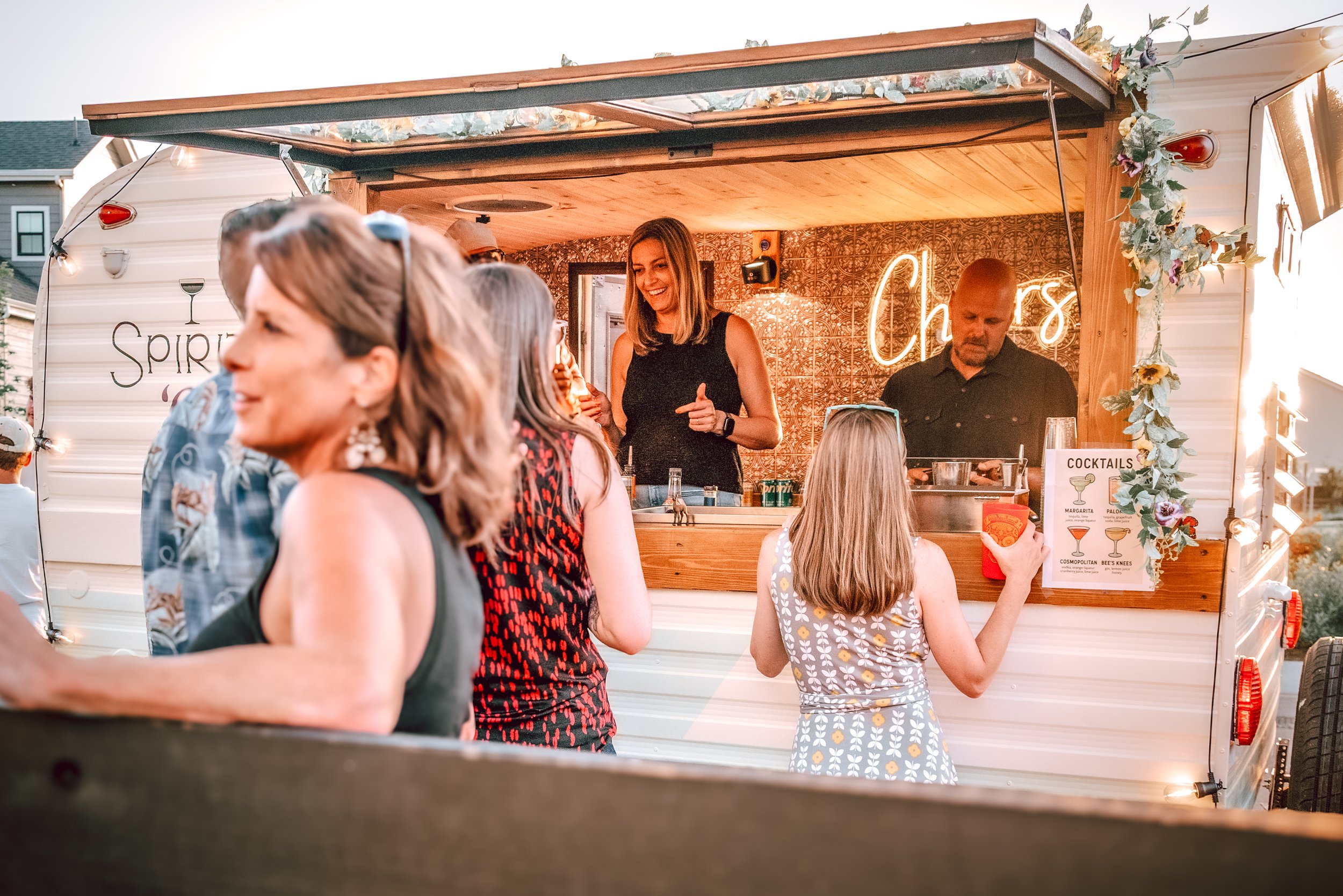 People socializing at a mobile bar with a neon sign that says 'Cheers' during sunset, decorated with flowers and greenery.